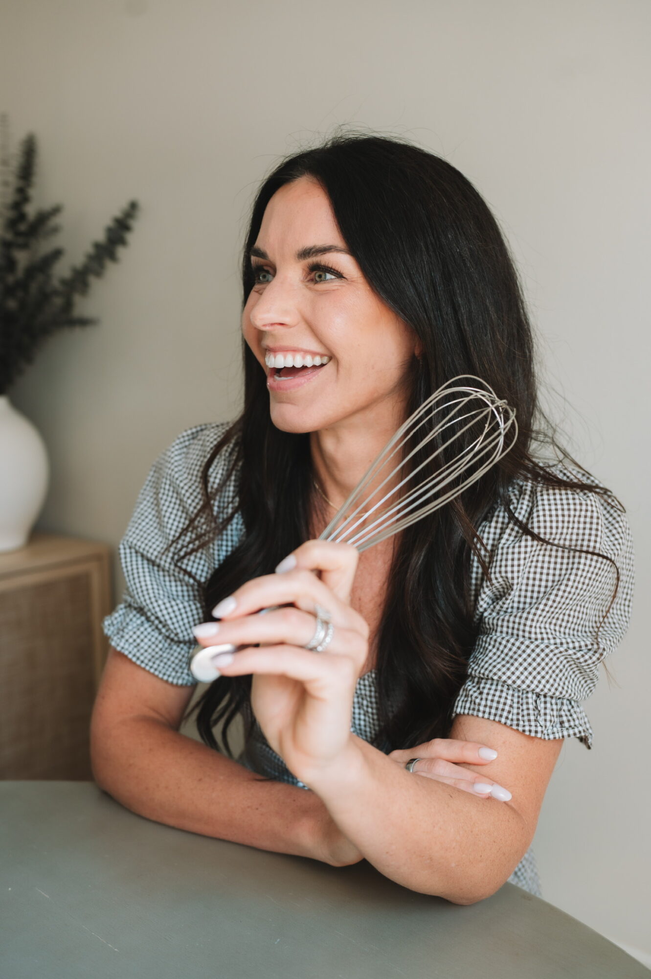 Woman with long dark hair smiling, holding a whisk, sitting at a table, in a room with a plant in the background.
