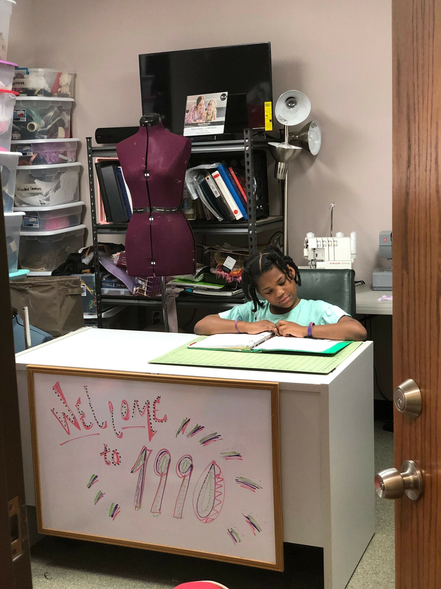 Person sitting at desk with computer and office supplies, framed handwritten note on desk, office environment with shelves and equipment.