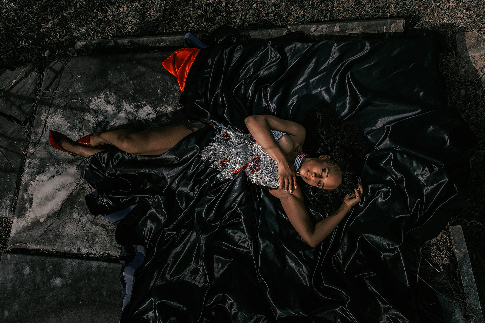 Woman lying on a black satin sheet outdoors, wearing a dress and red shoes, with her hand on her face.