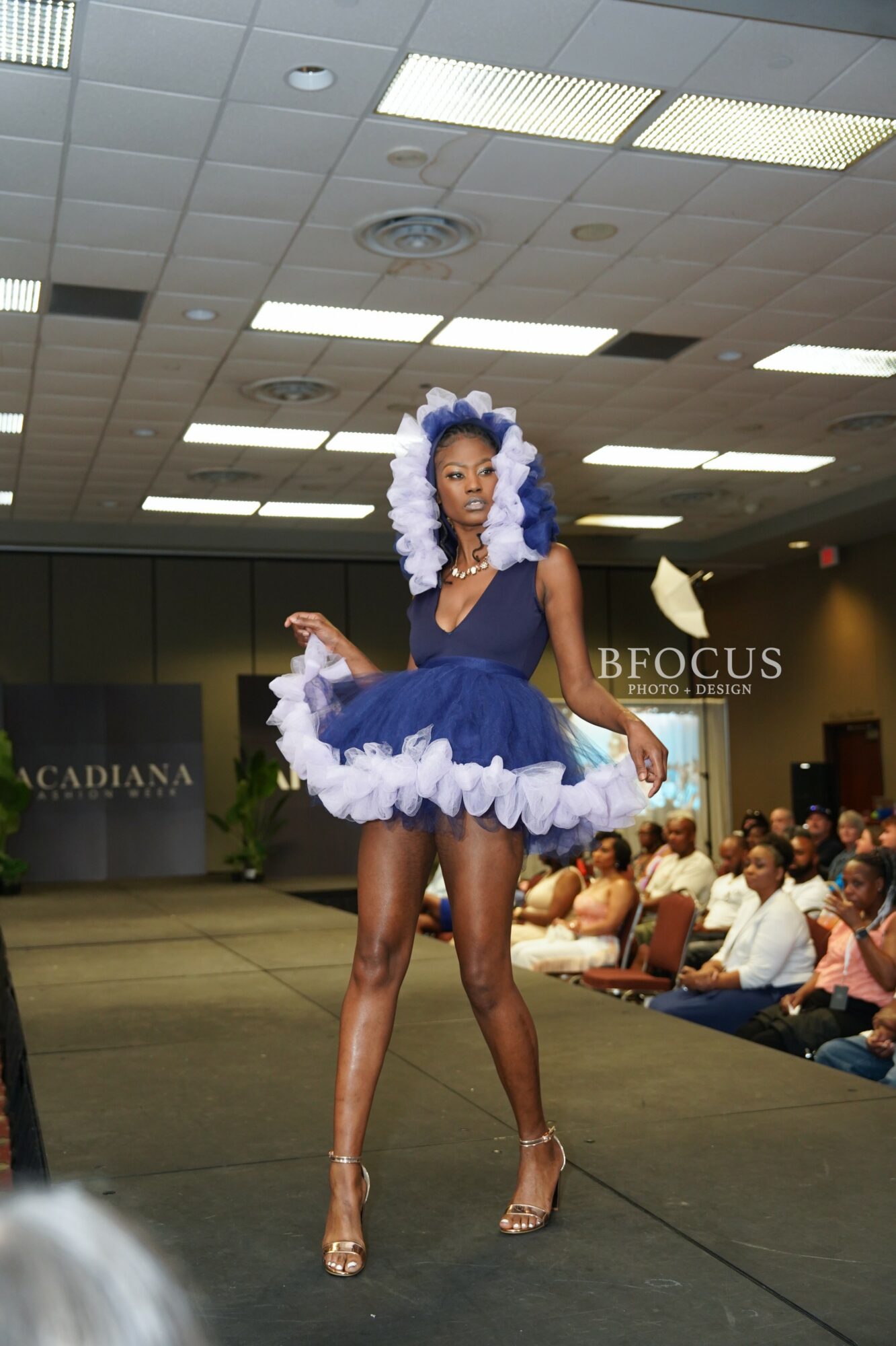 Model walking on runway in blue and white dress with large headpiece, audience seated on sides, ceiling lights overhead.