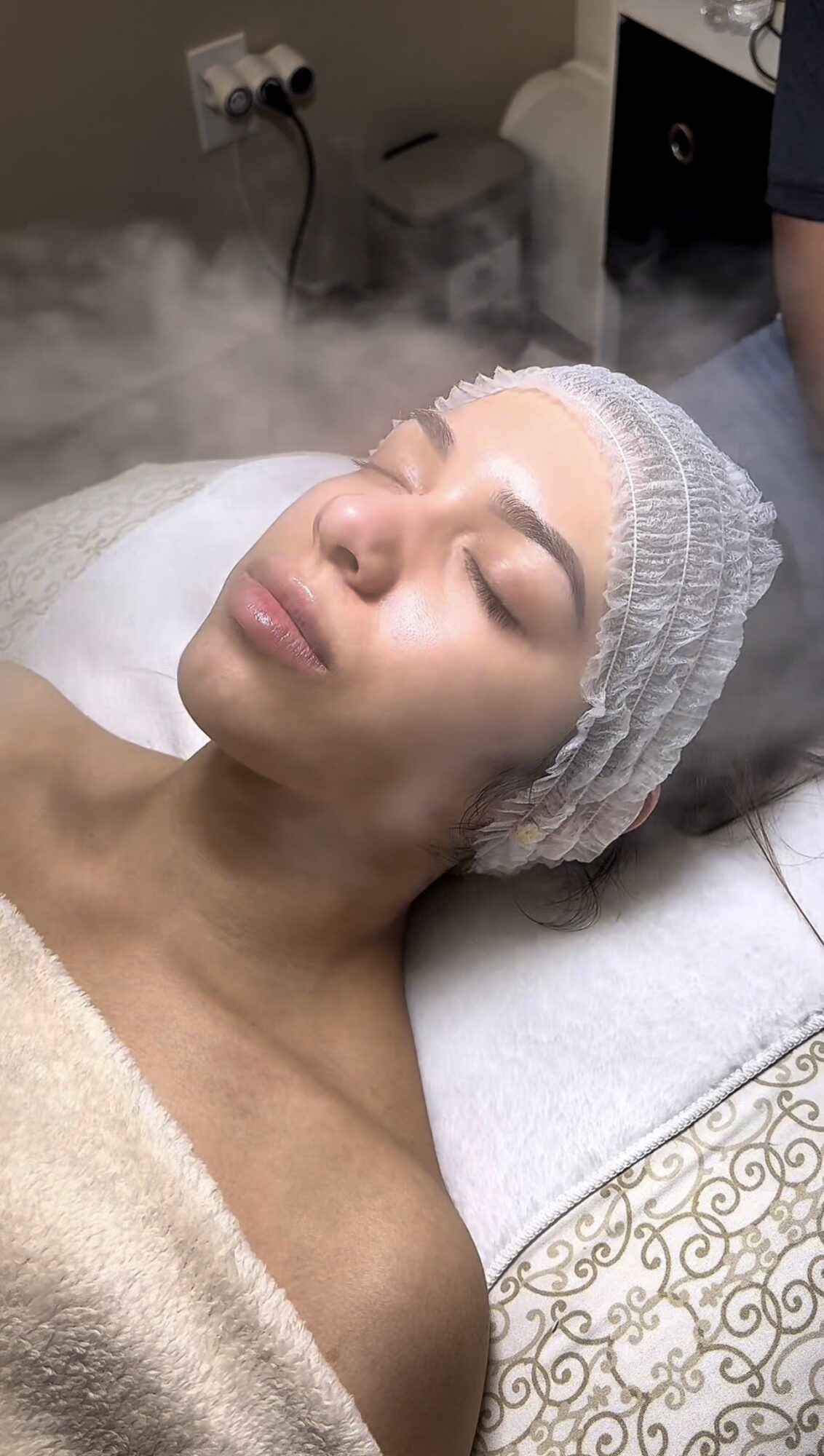 Woman with a hair cap receiving a facial treatment, lying with eyes closed, steam around her face, in a spa setting.