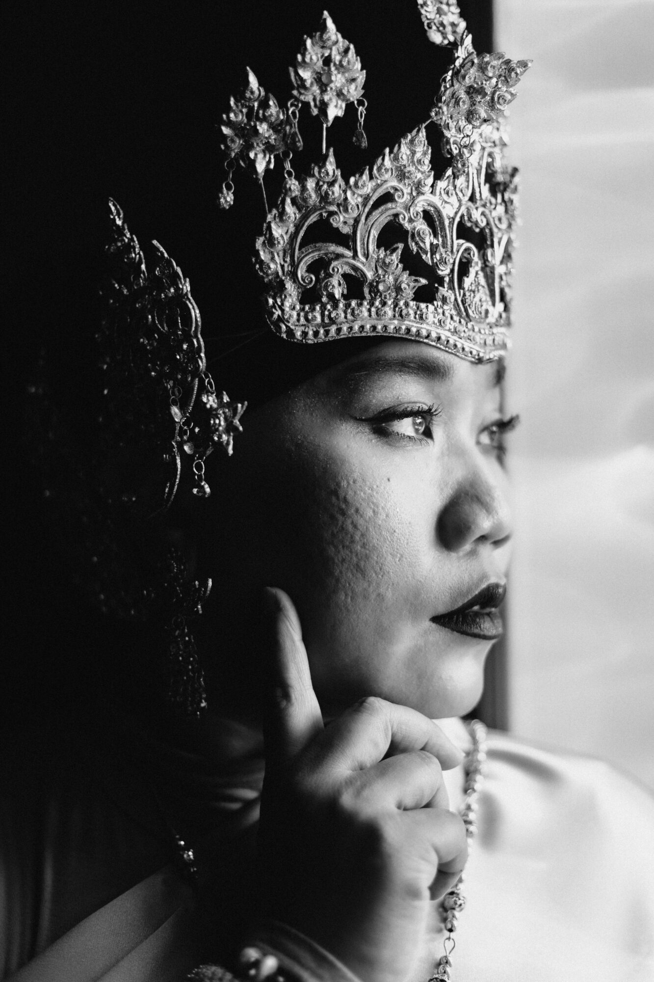 Woman wearing an ornate crown, looking out a window, with her hand near her face, in black and white.