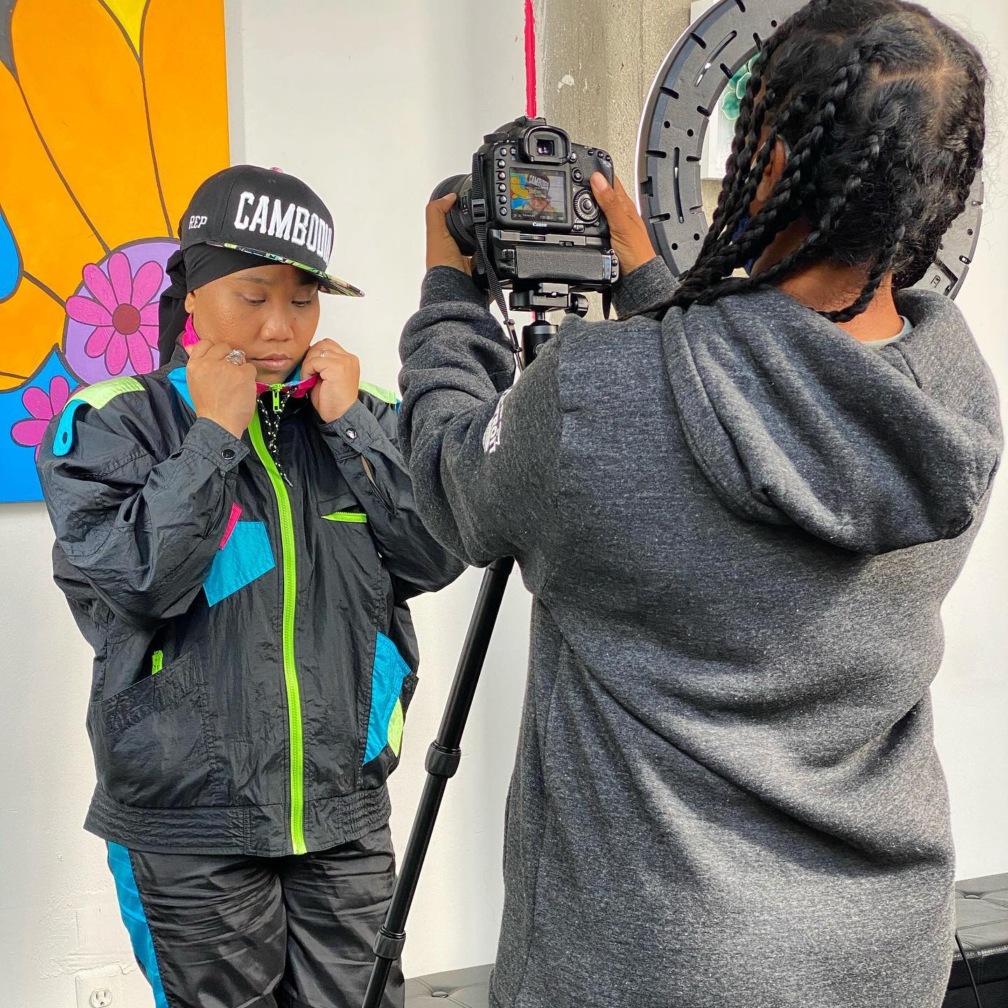 Child in black jacket with green accents and a black cap being photographed by a person with braided hair in a gray hoodie, indoors.
