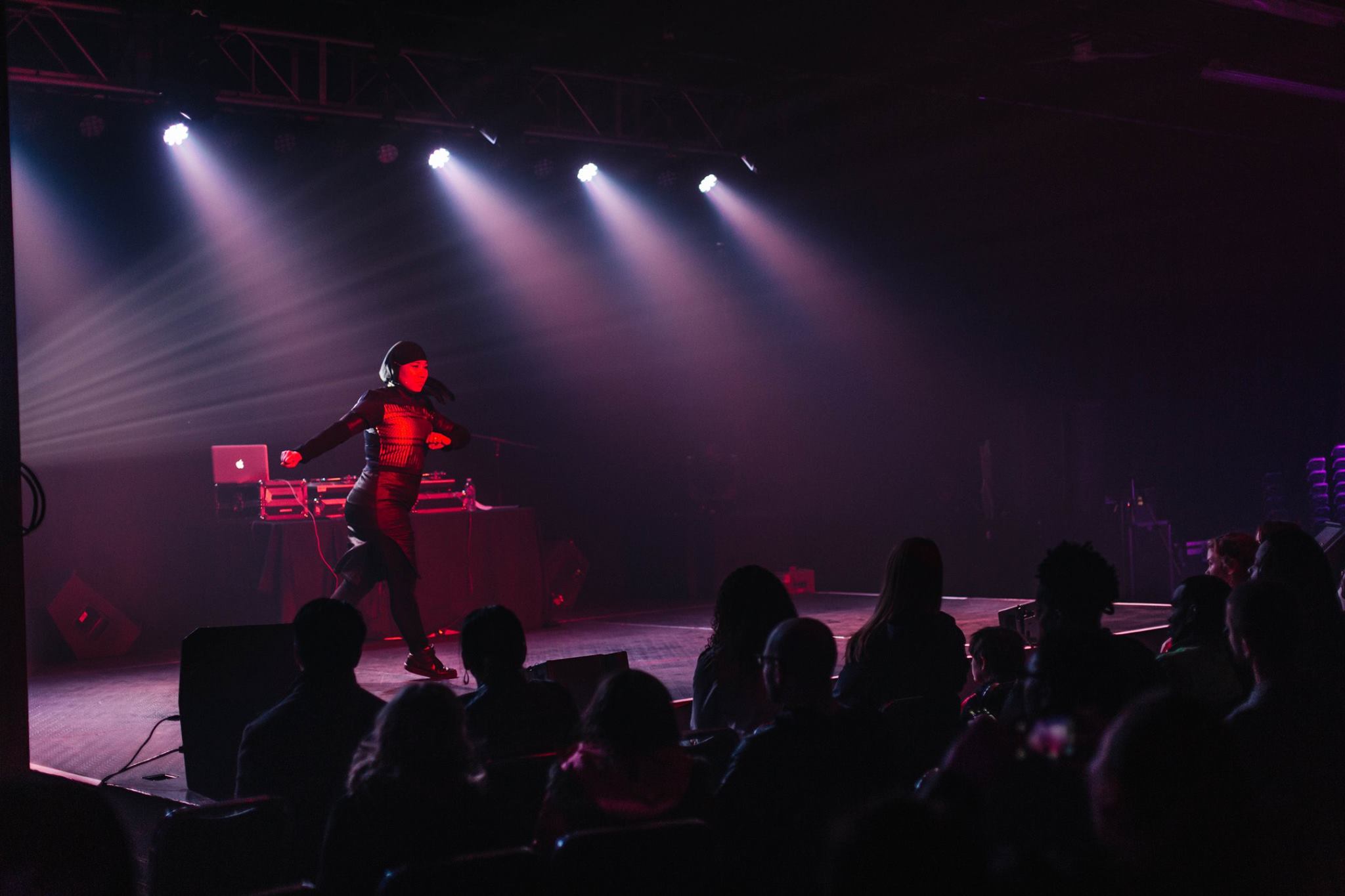 Performer on stage with DJ equipment, audience watching in a dark venue with purple lighting.