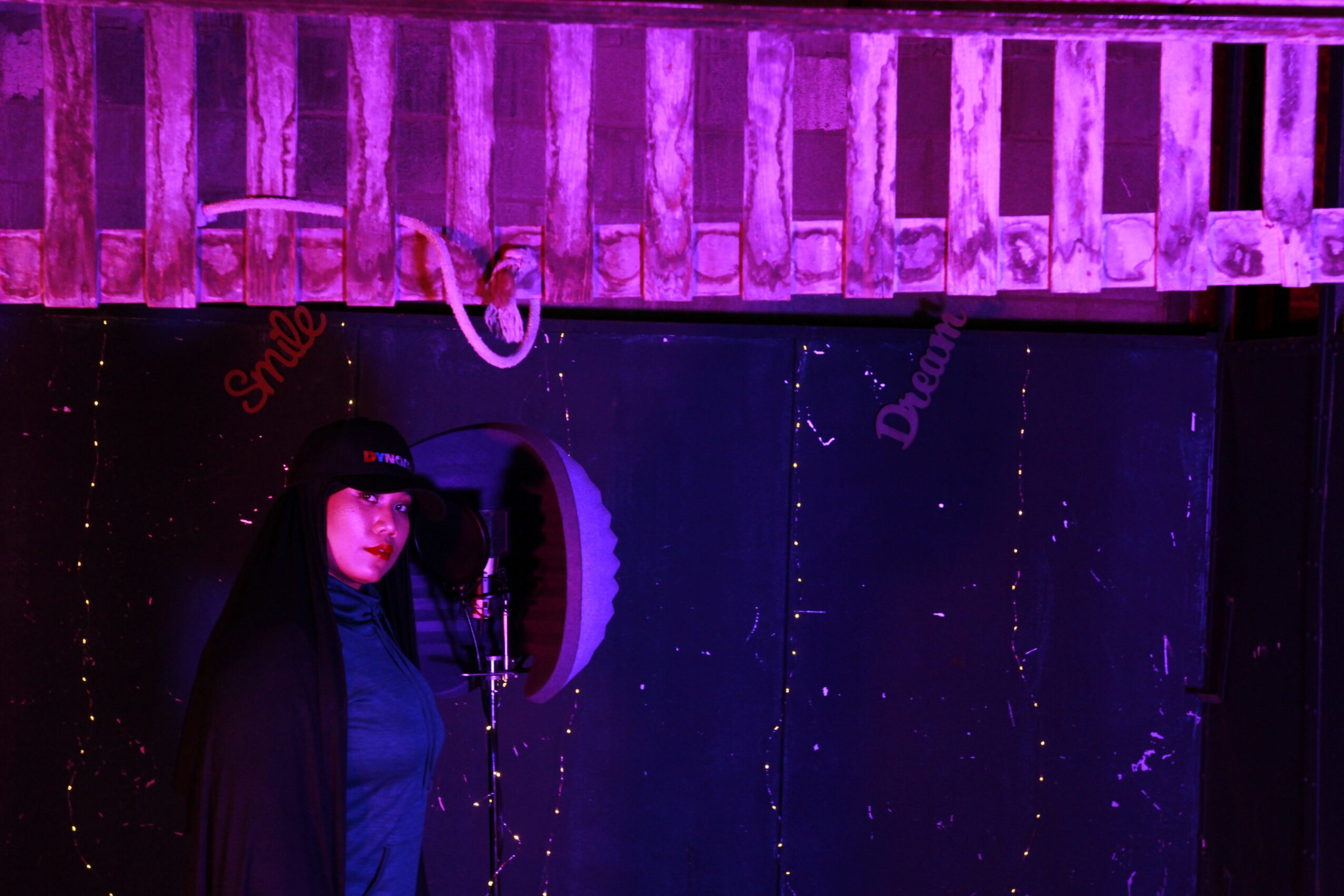 Person in dark clothing and hat standing in front of a microphone on a stage with purple lighting and a wooden railing.