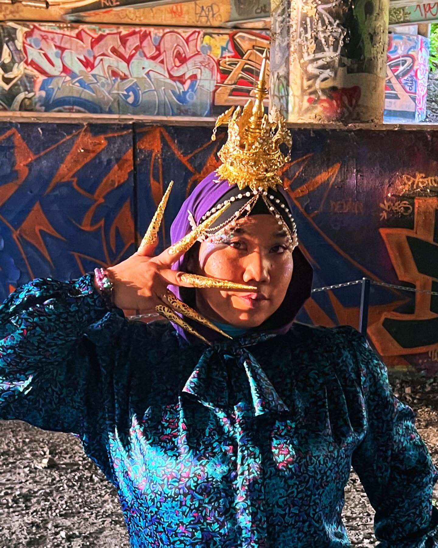 Person wearing a traditional costume with a golden headdress, posing with fingers near face, graffiti background.