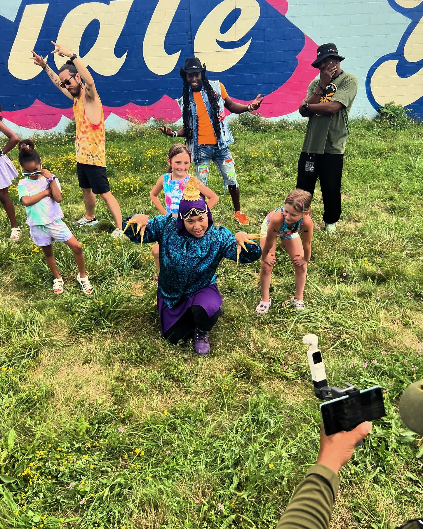 Group of children and adults outdoors on grass, some posing and others taking photos, with a colorful mural in background.