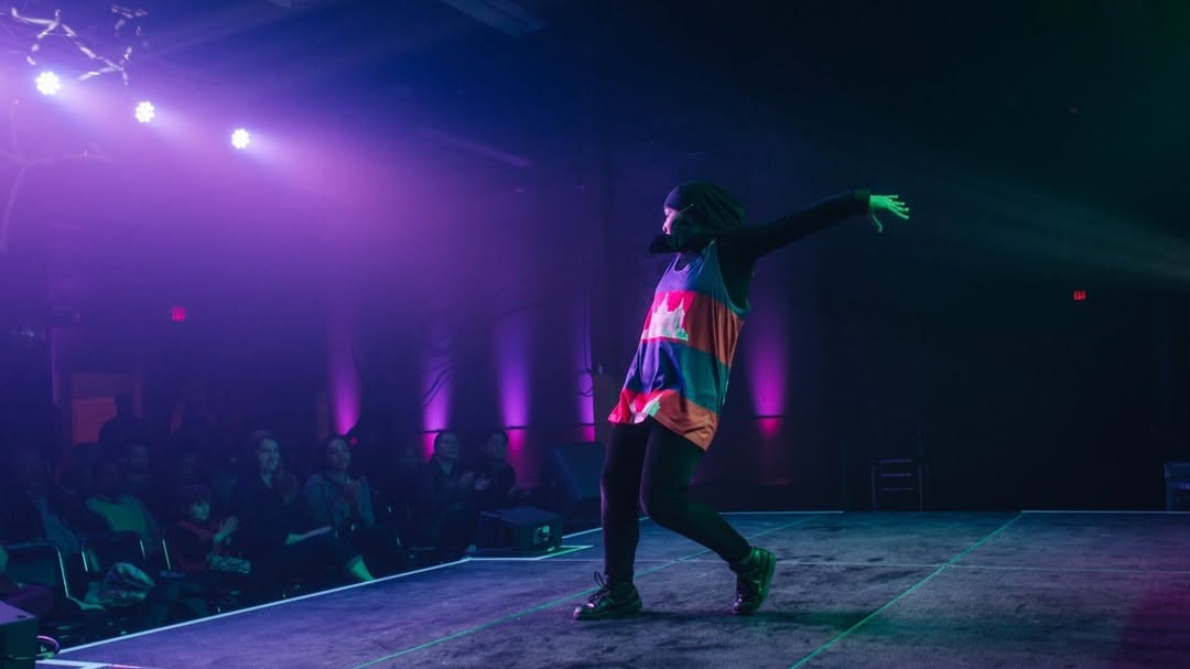 Performer on stage with colorful dress, arm extended, audience in background, purple and green lighting, dark venue.