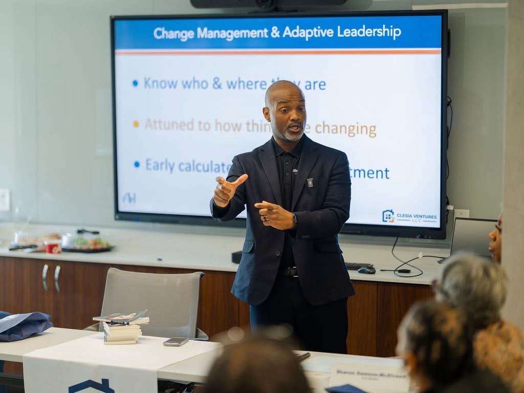 Man in dark suit giving presentation in front of a screen with text, audience seated in a conference room.