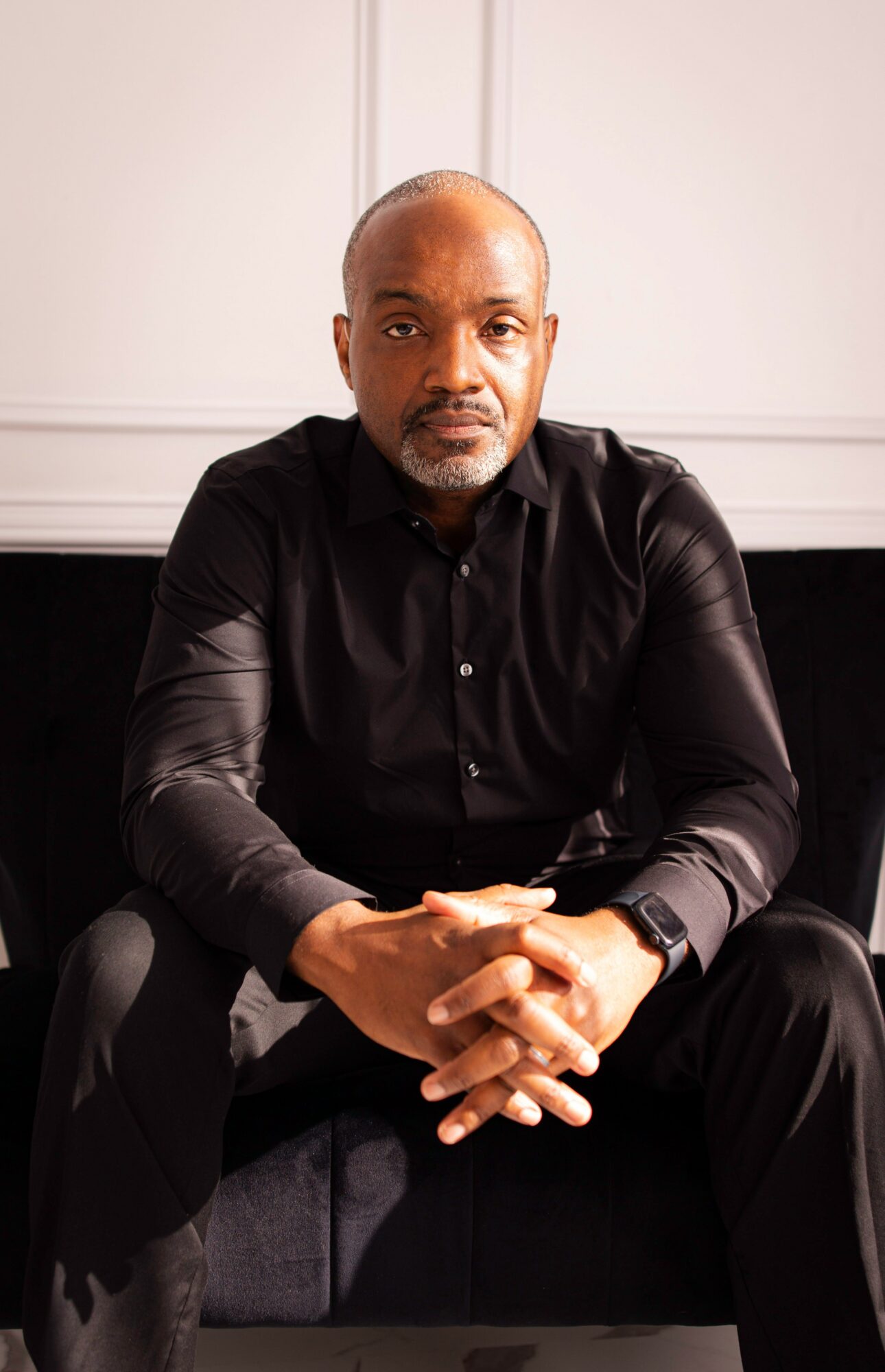 Man with a beard and bald head sitting with hands clasped, wearing a black shirt, in a room with white walls.