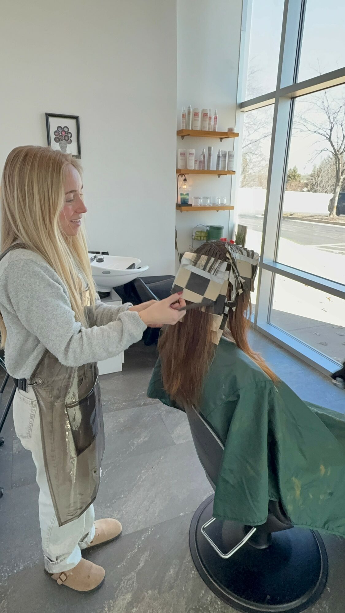 A woman with long hair is sitting in a salon chair, getting her hair colored by a stylist holding a color palette, near a large window.