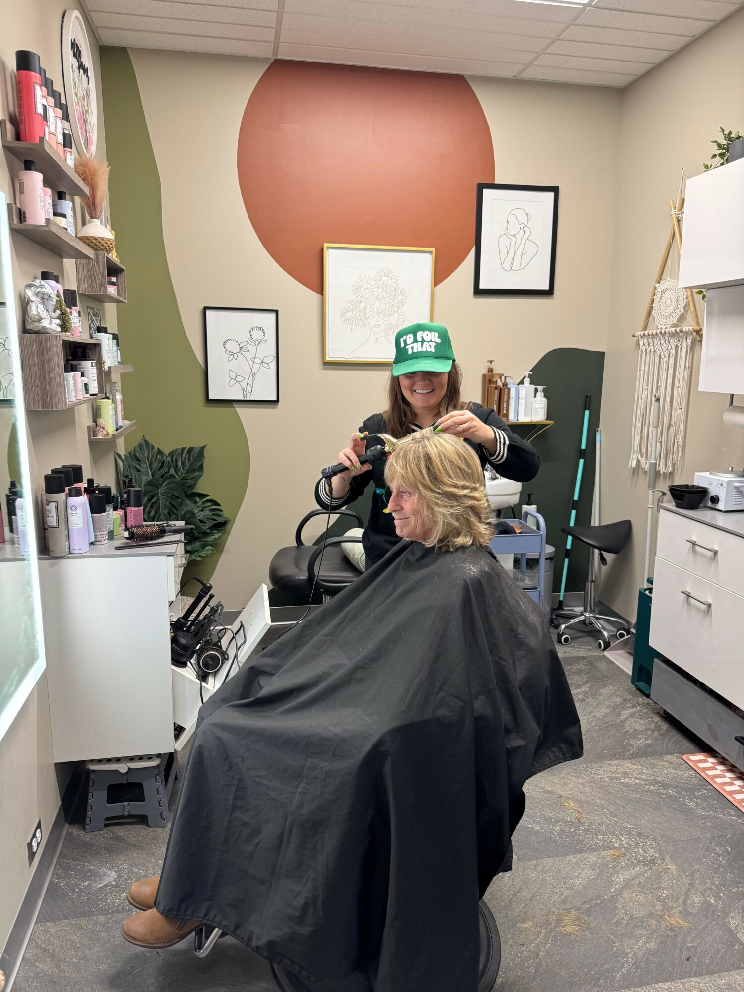 A person with blonde hair sits in a salon chair, wearing a black cape, getting a haircut. The salon has framed artwork and shelves with products.