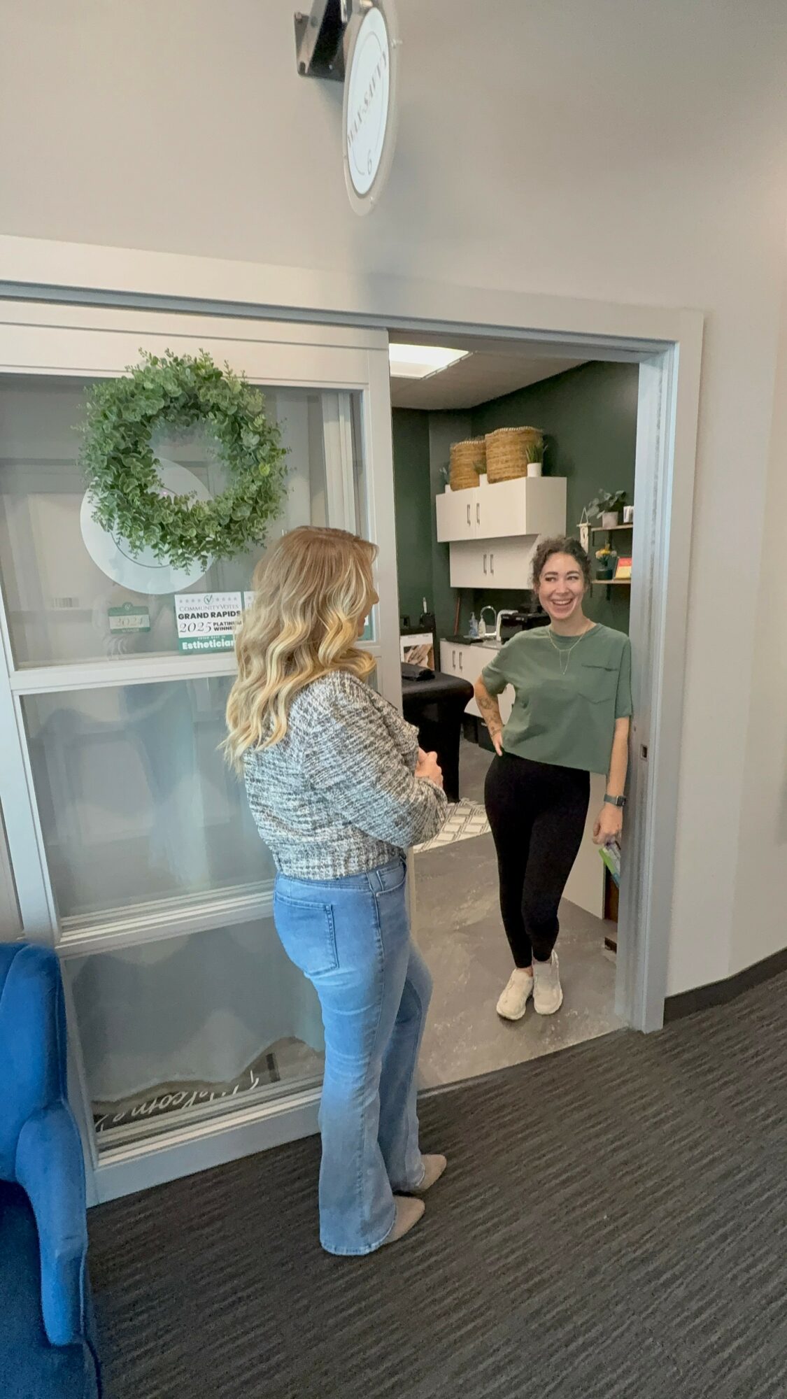 Two women standing and talking in an office hallway, one facing the camera and the other with her back to it, near a glass door with a wreath.