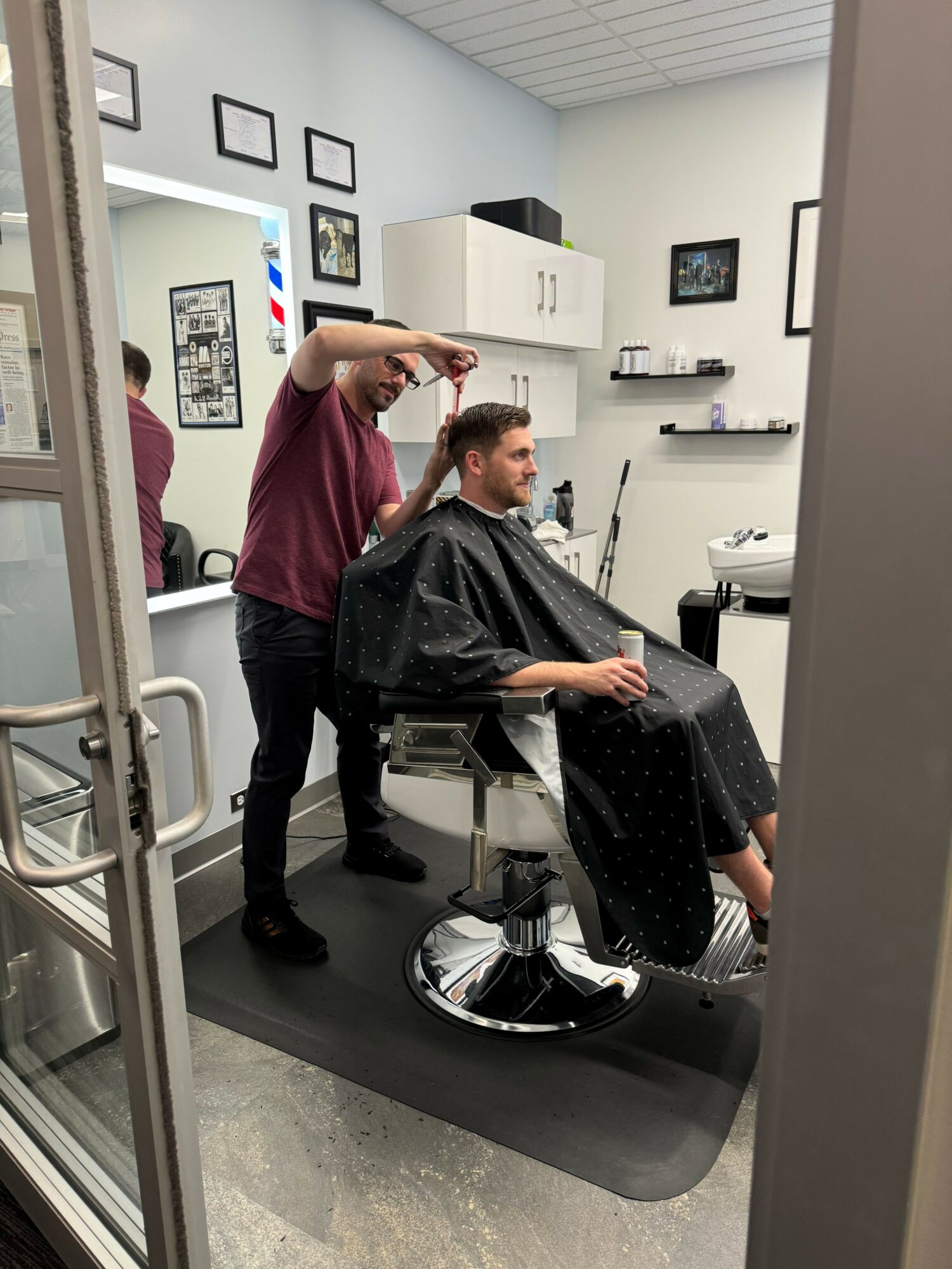 Two people in a hair salon, one sitting in a chair and the other standing behind, with salon equipment in the background.