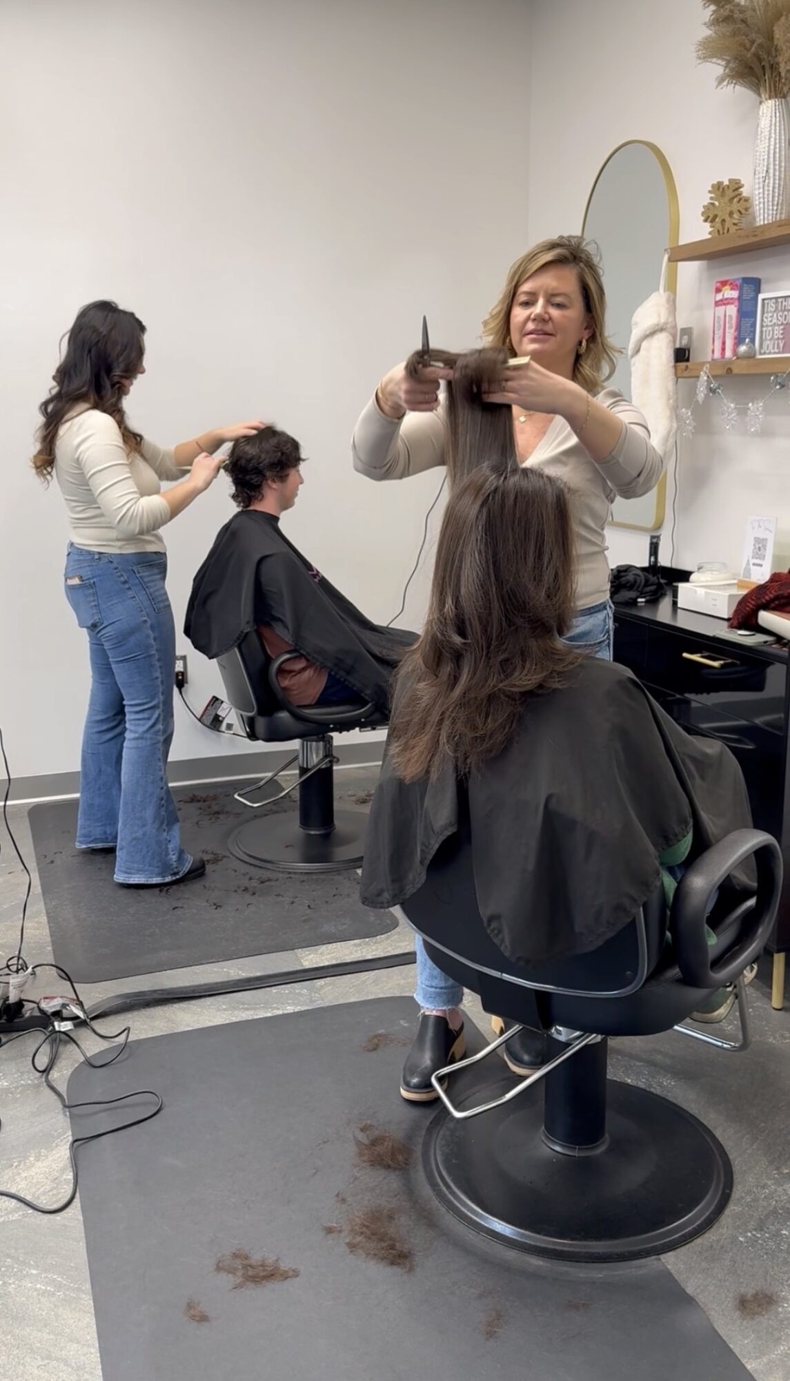 A woman takes a photo of a person getting a haircut in a salon, with another person standing nearby. Two people are seated in salon chairs.