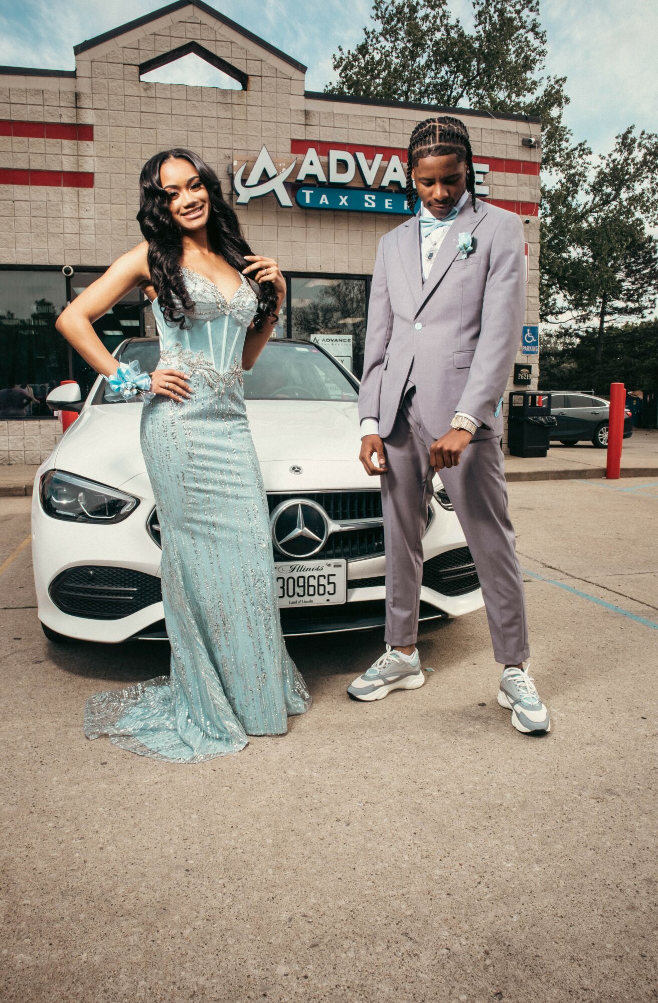Two young people in formal attire stand in front of a white Mercedes-Benz car outside a building with a sign reading 'ADVANCE TAX SERVICE'.