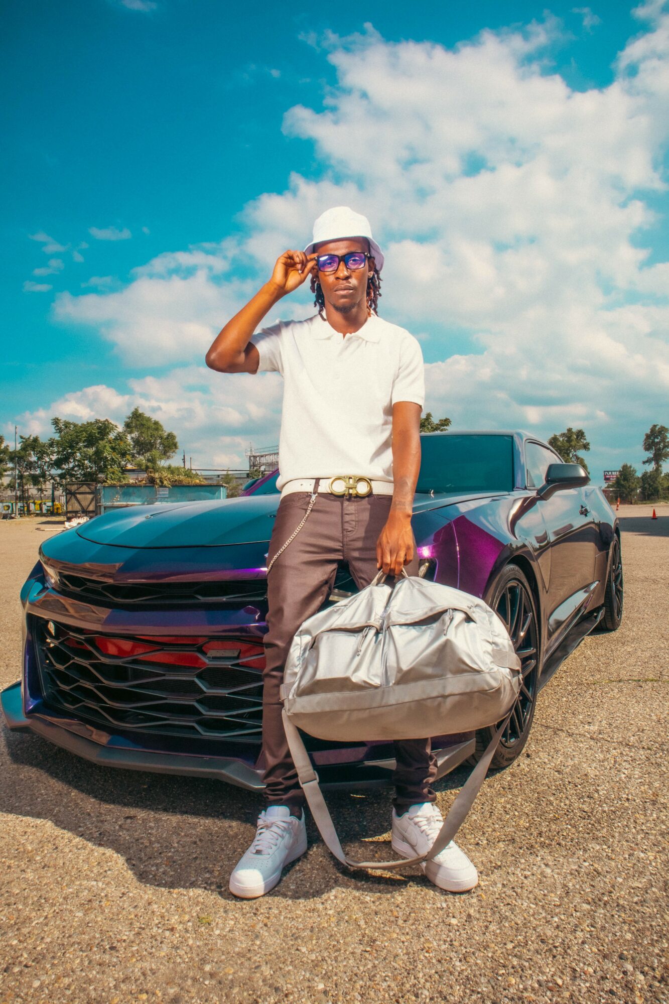 Person standing in front of a black sports car, holding a large bag, wearing a white hat and glasses, outdoors under blue sky