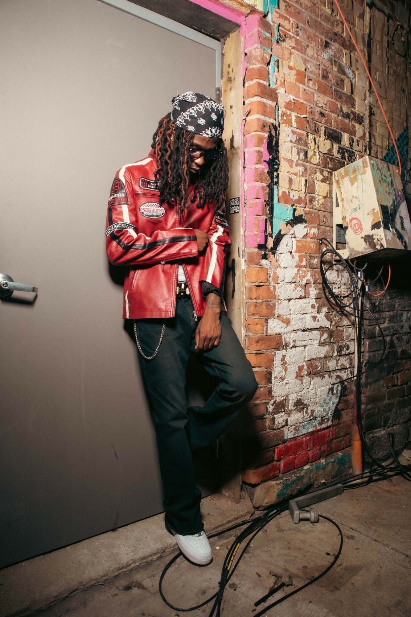 Person with dreadlocks wearing a red leather jacket, black pants, and white shoes leaning against a brick wall.