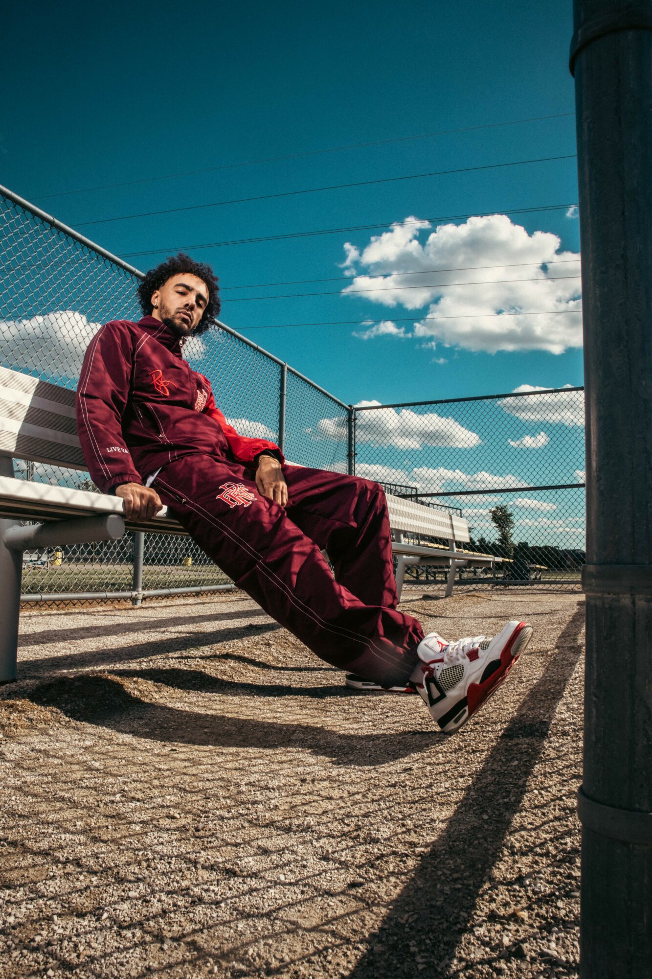 Person sitting on a bench outdoors, wearing a red outfit, with a blue sky and clouds in the background.