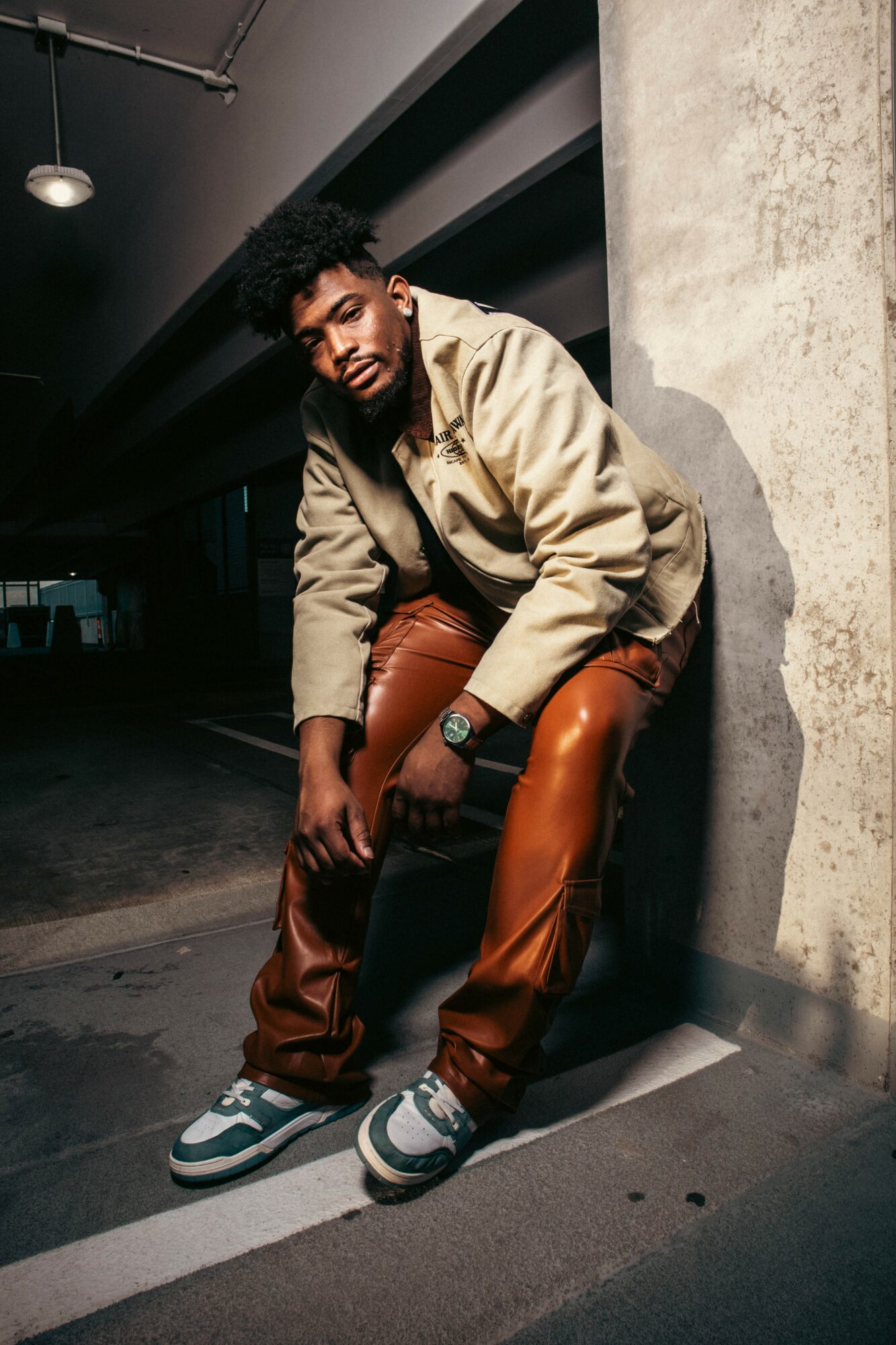 Person with curly hair and beard sitting against a concrete wall in an urban setting, wearing a beige jacket and brown pants.