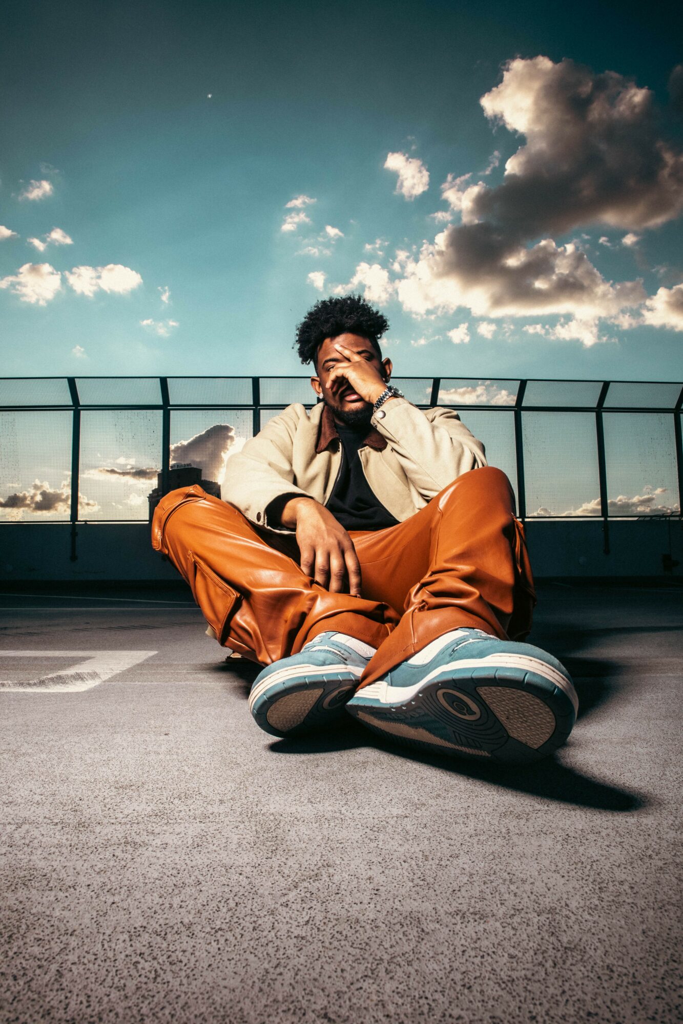 Person sitting on ground with legs crossed, hands covering face, sky with clouds in background.