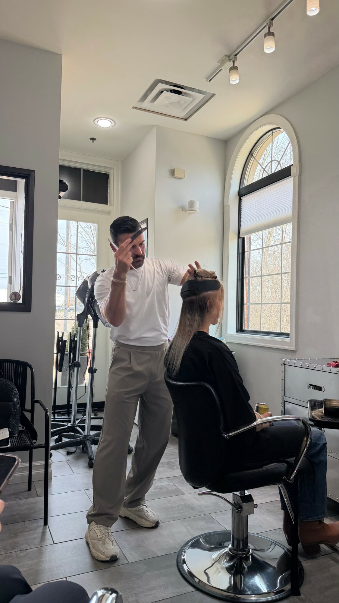 A hairstylist stands behind a seated woman in a salon, working on her hair near large arched windows.