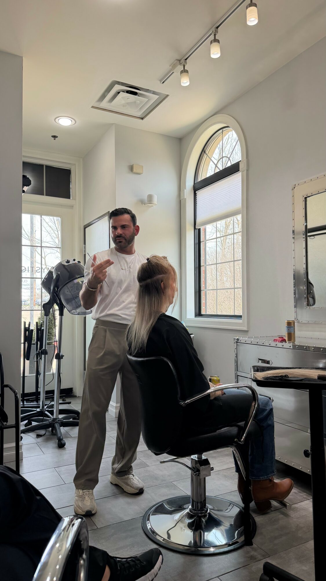 A hairstylist stands next to a seated woman in a salon, with large windows and bright lighting in the background.