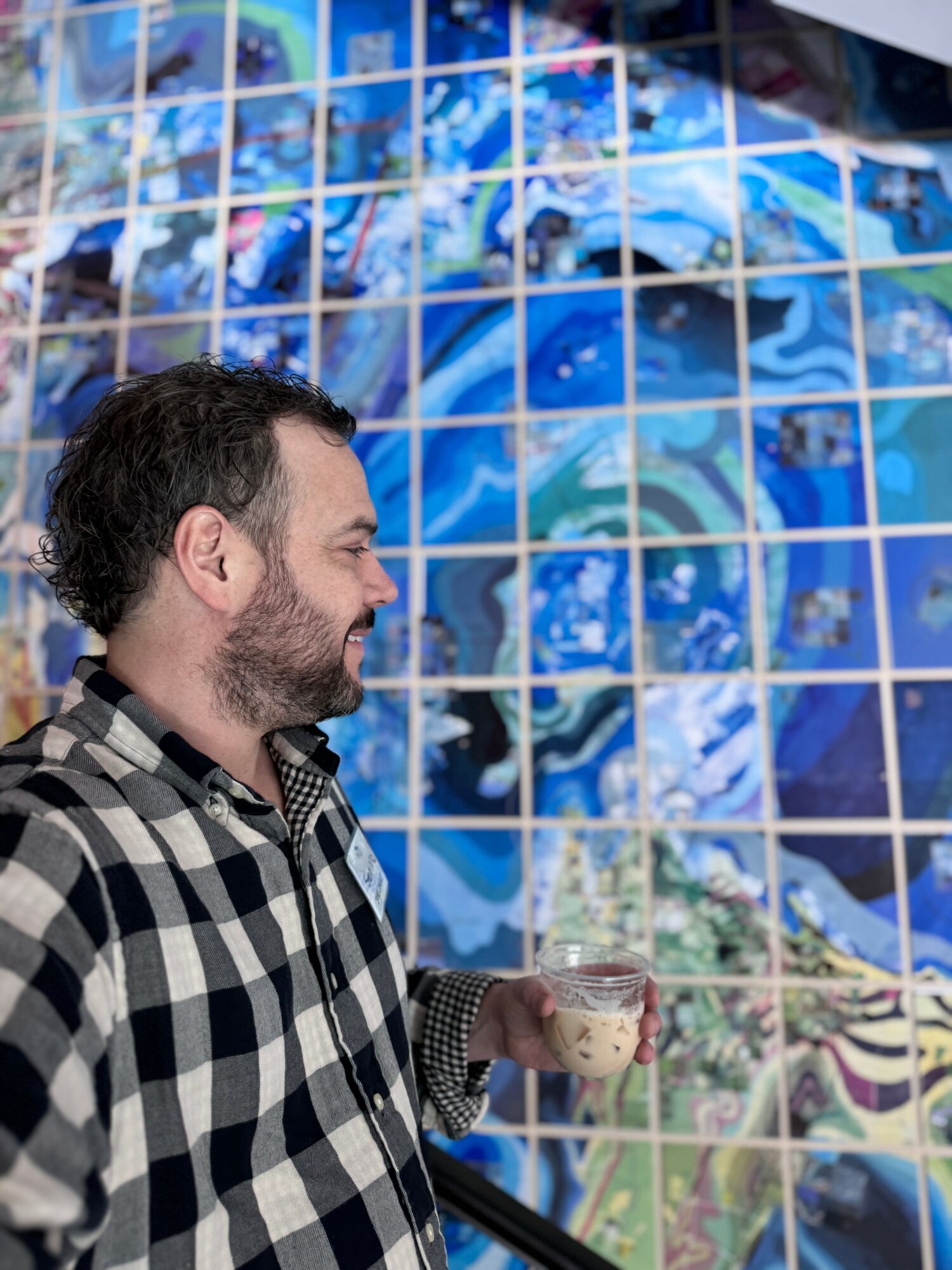 Man with dark hair and beard in checkered shirt holding a drink, standing in front of a colorful mosaic wall.