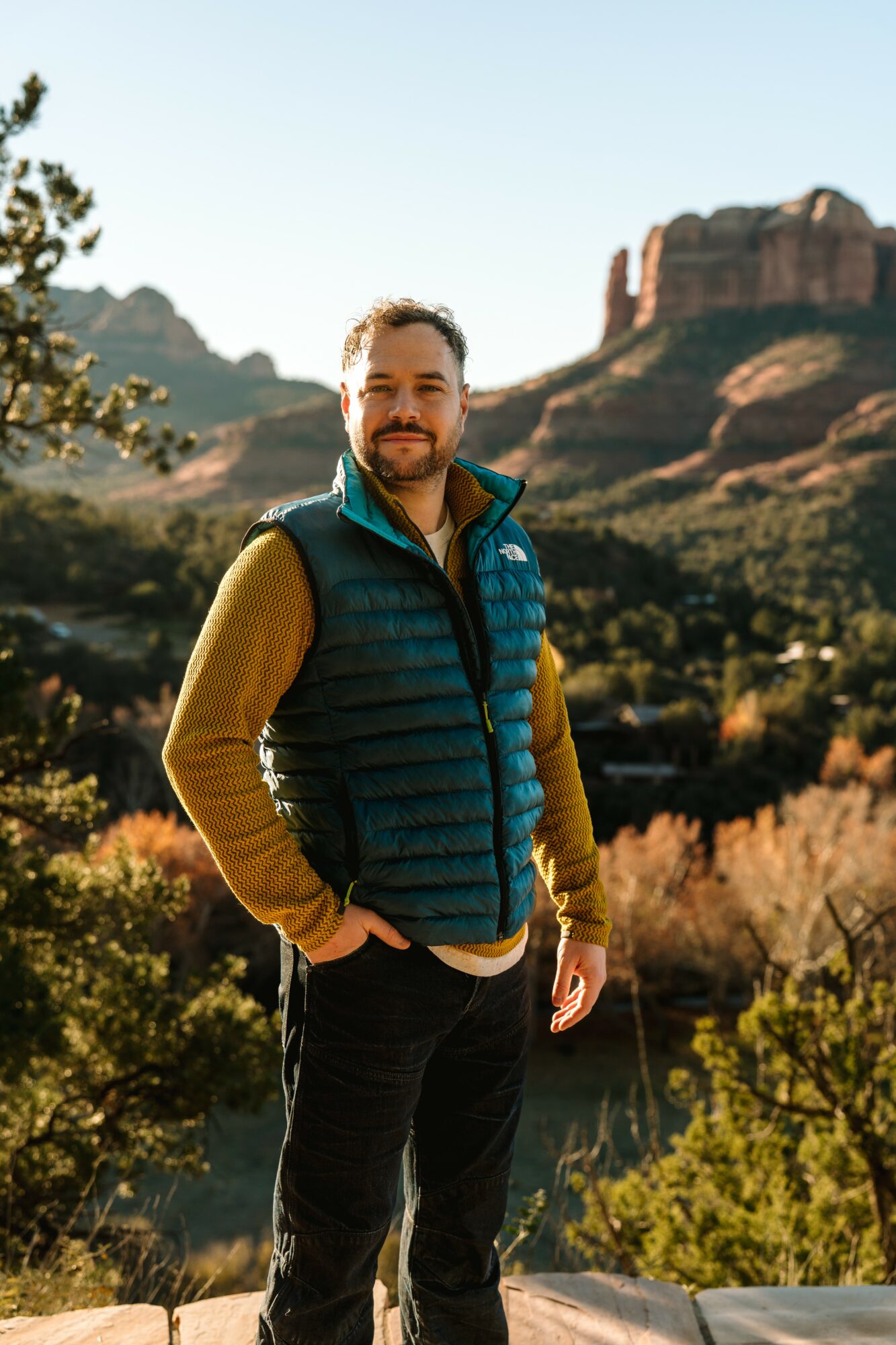 Man standing outdoors with mountains and trees in background, smiling, wearing a blue vest and yellow sleeves, hands in pockets.