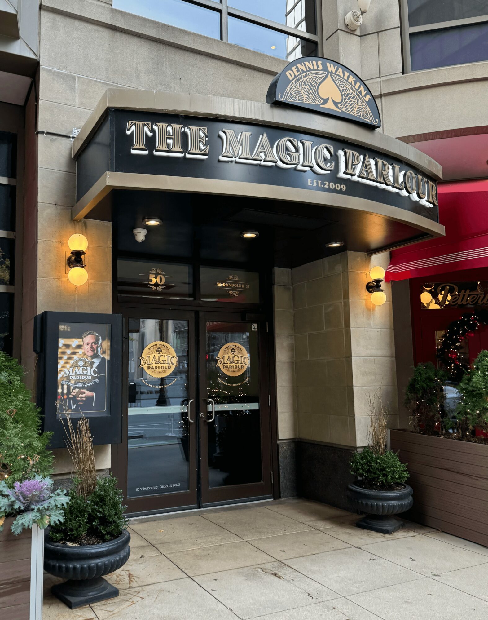 Entrance to The Magic Castle with glass doors, potted plants, and a black awning with gold lettering, outside a building.