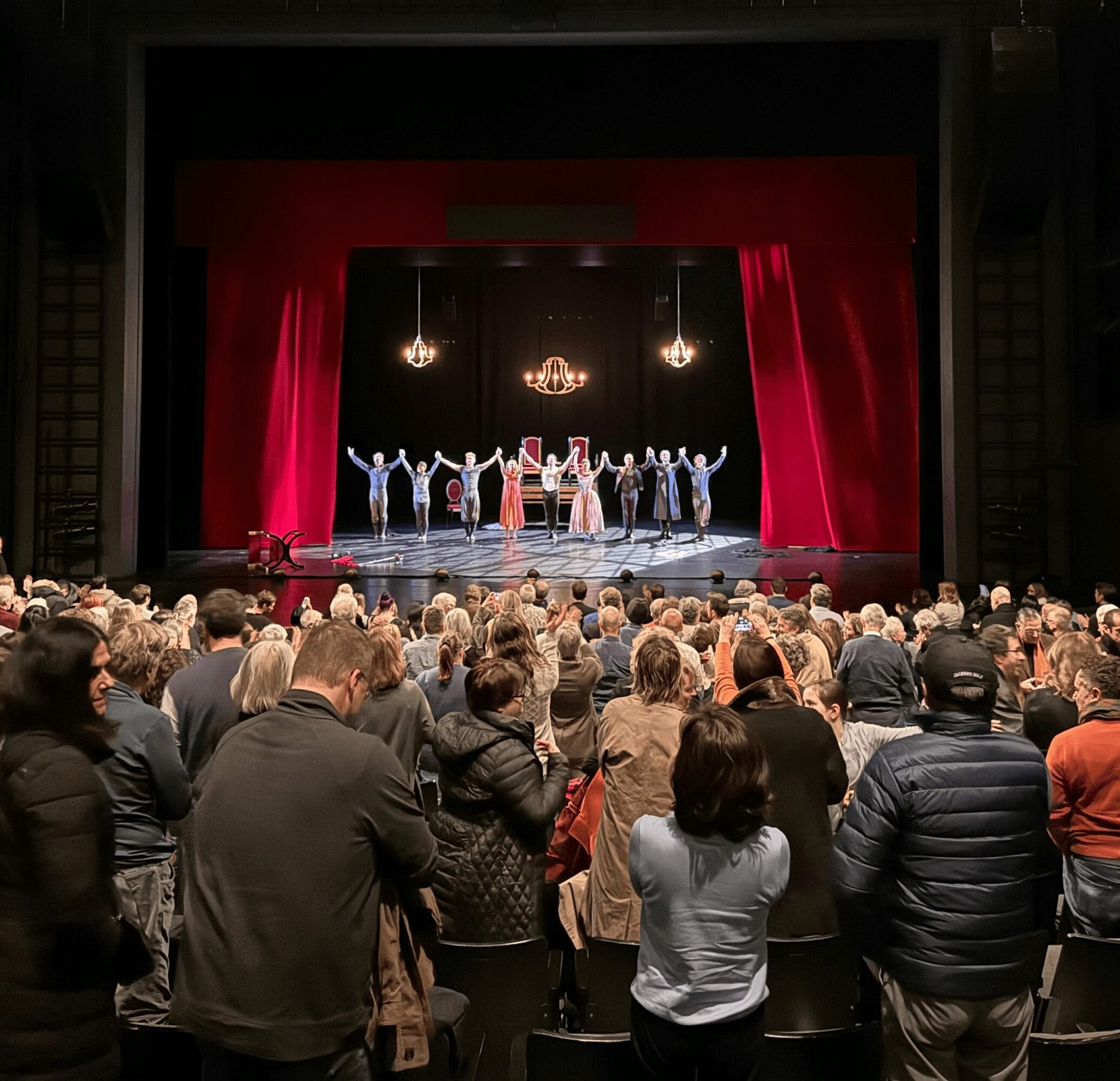 Audience watching a stage performance with dancers and a chandelier, red curtains framing the stage, in a theater setting.