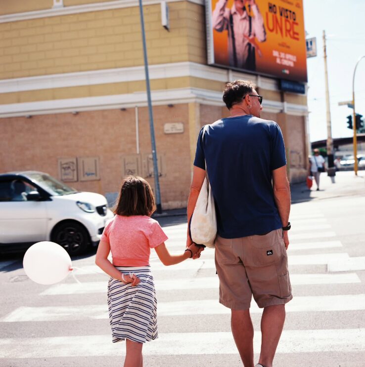 A man and a girl holding hands crossing a street on a sunny day, with cars and a billboard in the background.