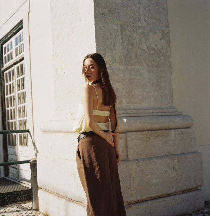 Young woman with long hair standing outdoors near a stone building, sunlight on her face and body, looking over her shoulder.