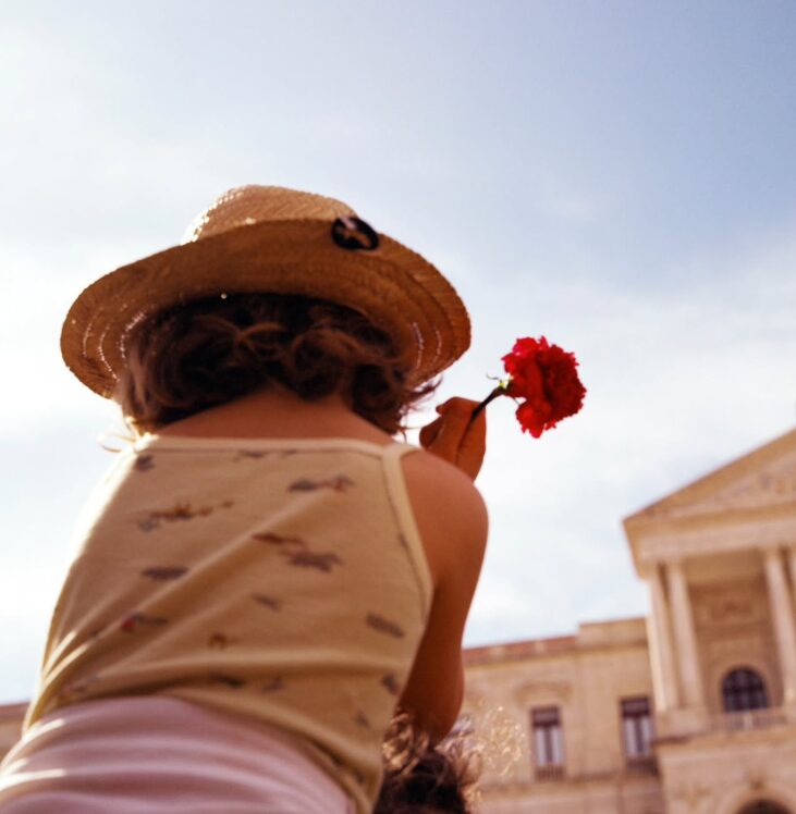 Child wearing a straw hat holding a red flower, facing a building with classical architecture, sky in background.
