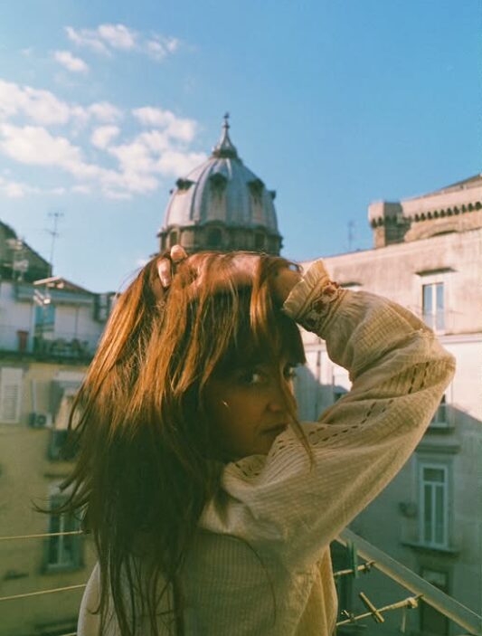 Woman with long hair and beige clothing on a balcony, with a domed building and blue sky in background.