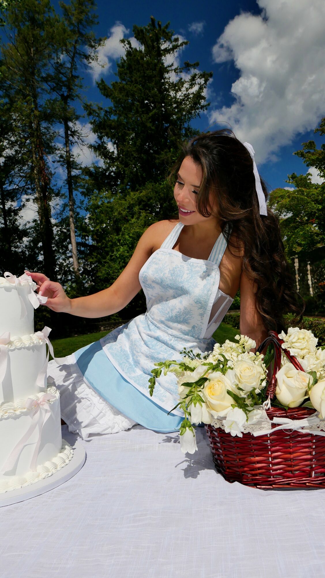 Woman in white apron with long hair, outdoors, with trees and sky, next to a cake and flowers in a basket.