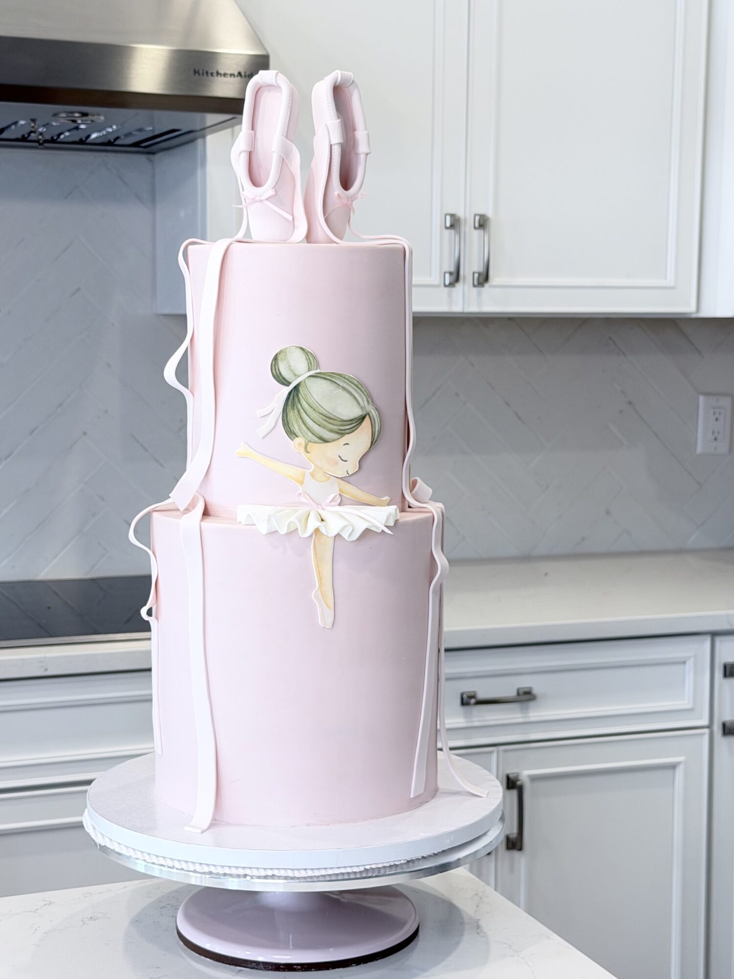 Pink cake with a ballerina figure, two ballet shoes on top, on a white cake stand in a kitchen.