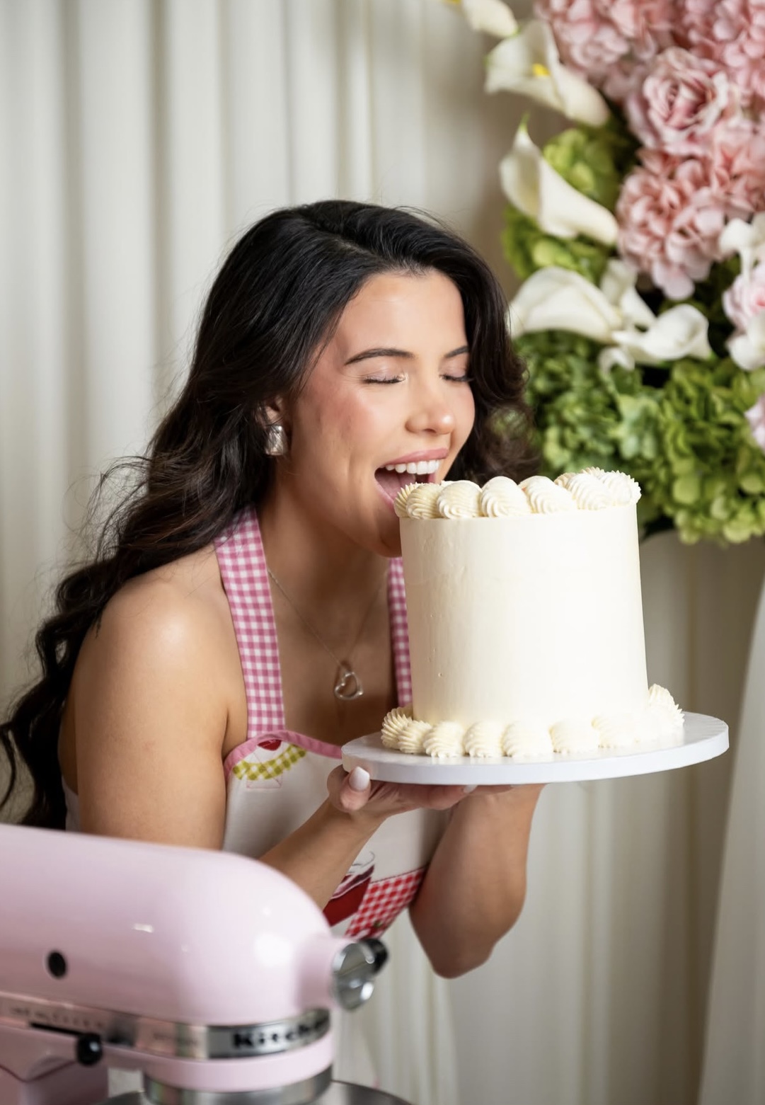 Young woman with long dark hair holding a birthday cake with white frosting, smiling, with pink and white flowers in background.
