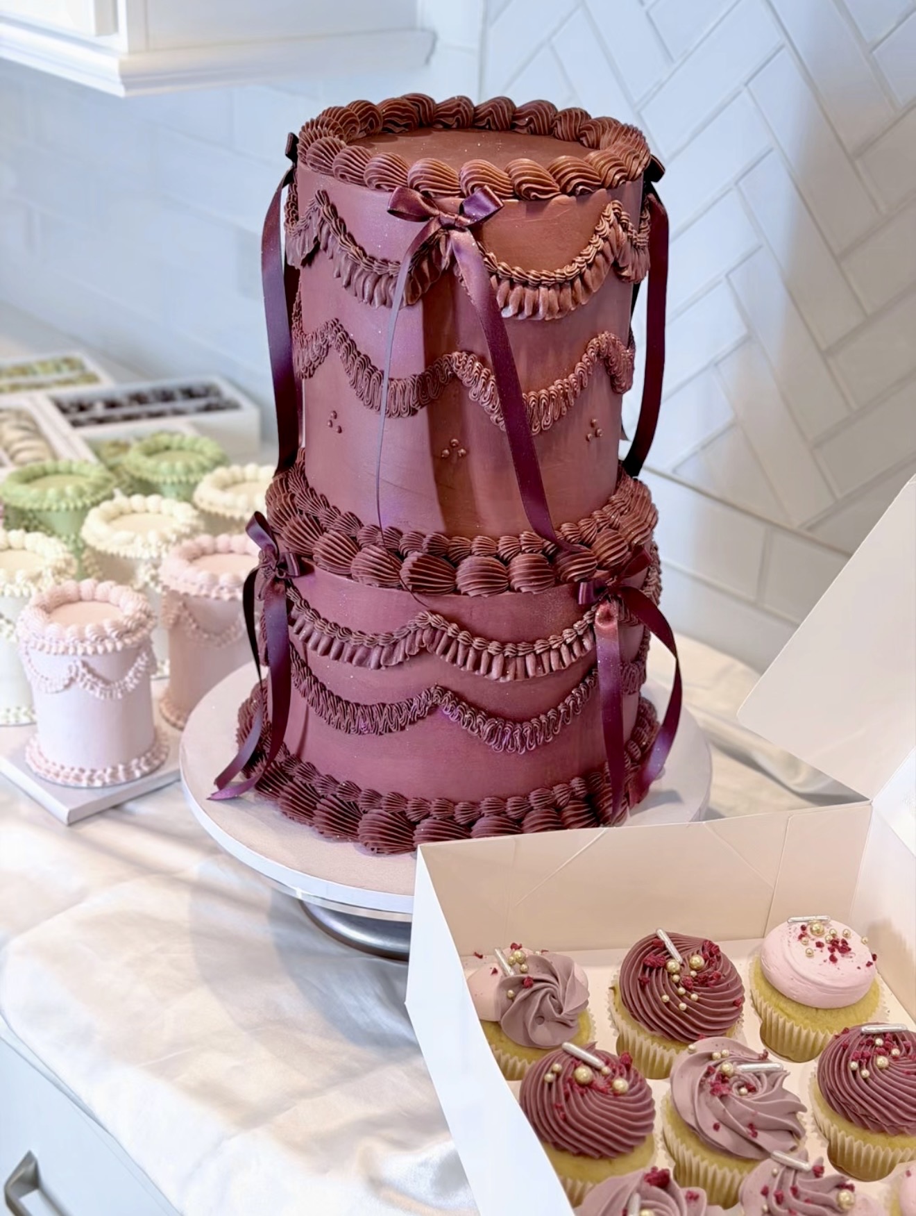 Two-tiered chocolate cake with decorative ribbons and piping, surrounded by cupcakes with pink and purple frosting, on a table.