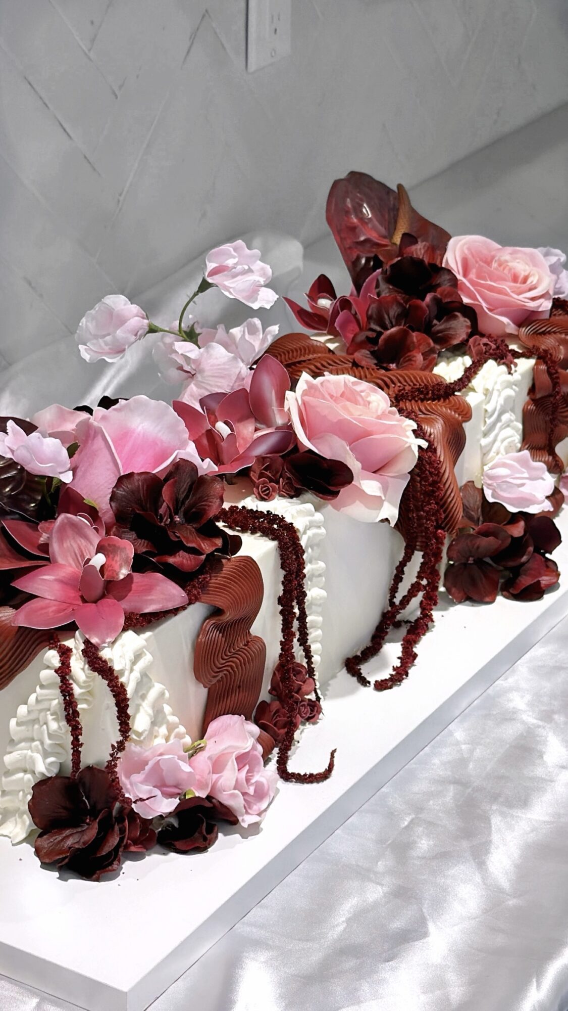 Decorative cake with pink and dark red flowers, draped with red strings, on a white surface against a gray wall.