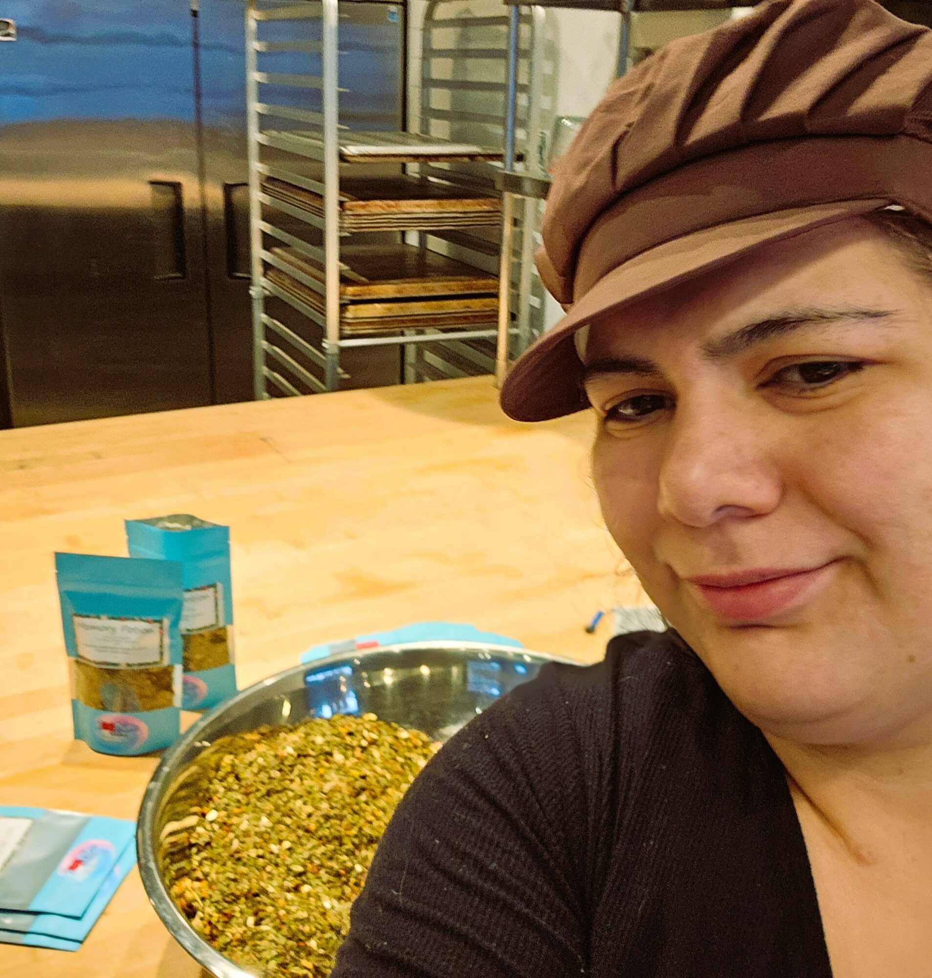 Person wearing a brown cap and black shirt, smiling, in a kitchen with baking racks and a wooden counter, bowl of food in foreground.