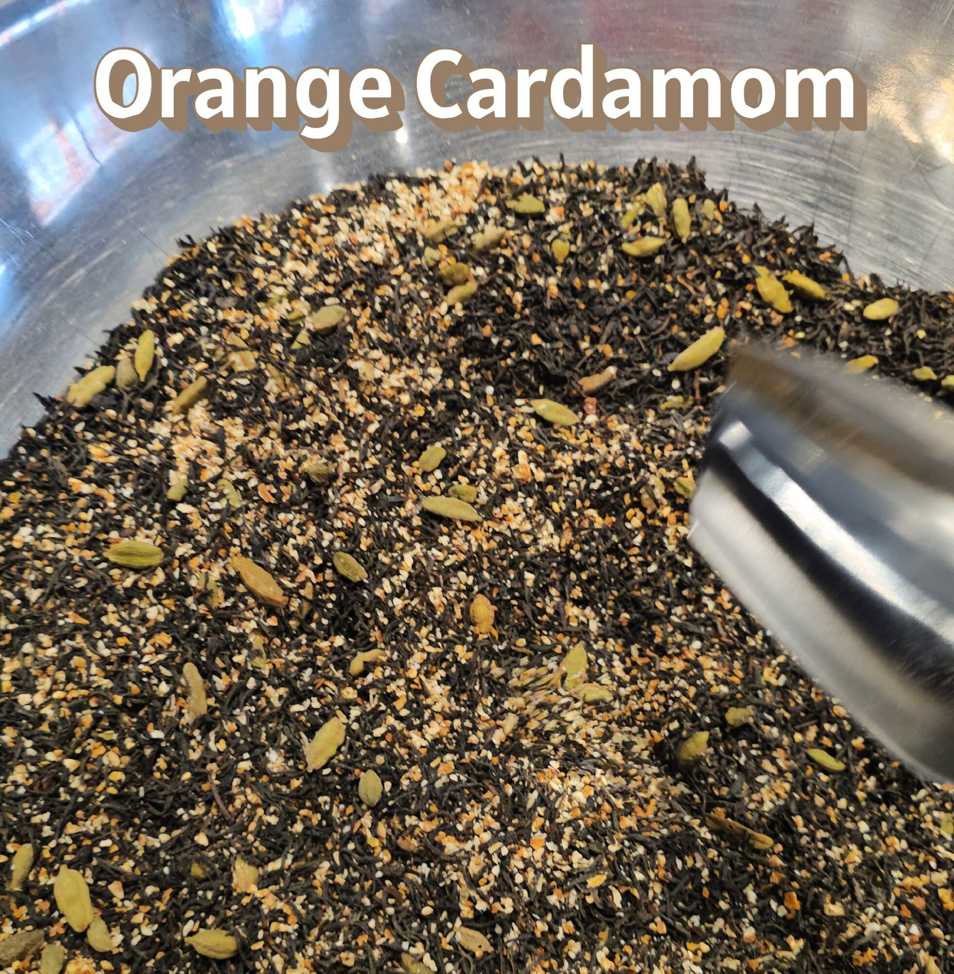 Close-up of a mixture of black seeds, chopped nuts, and orange zest in a metal bowl, labeled 'Orange Cardamom'.