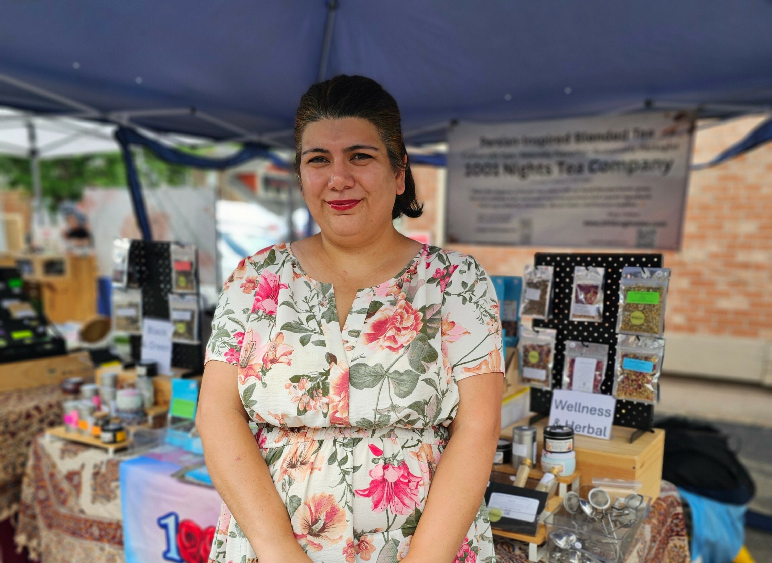 Woman standing in front of a booth at an outdoor market, smiling, wearing a floral dress.