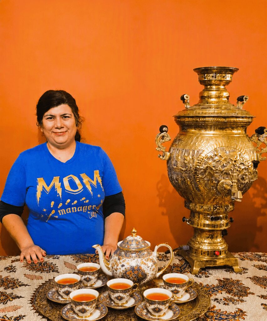 Woman in blue shirt standing behind a table with tea set and large ornate gold samovar against orange background.