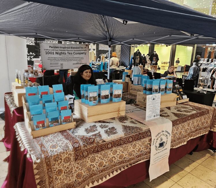Woman sitting behind a table with blue packaged products at an indoor market stall, with other stalls and people in the background.