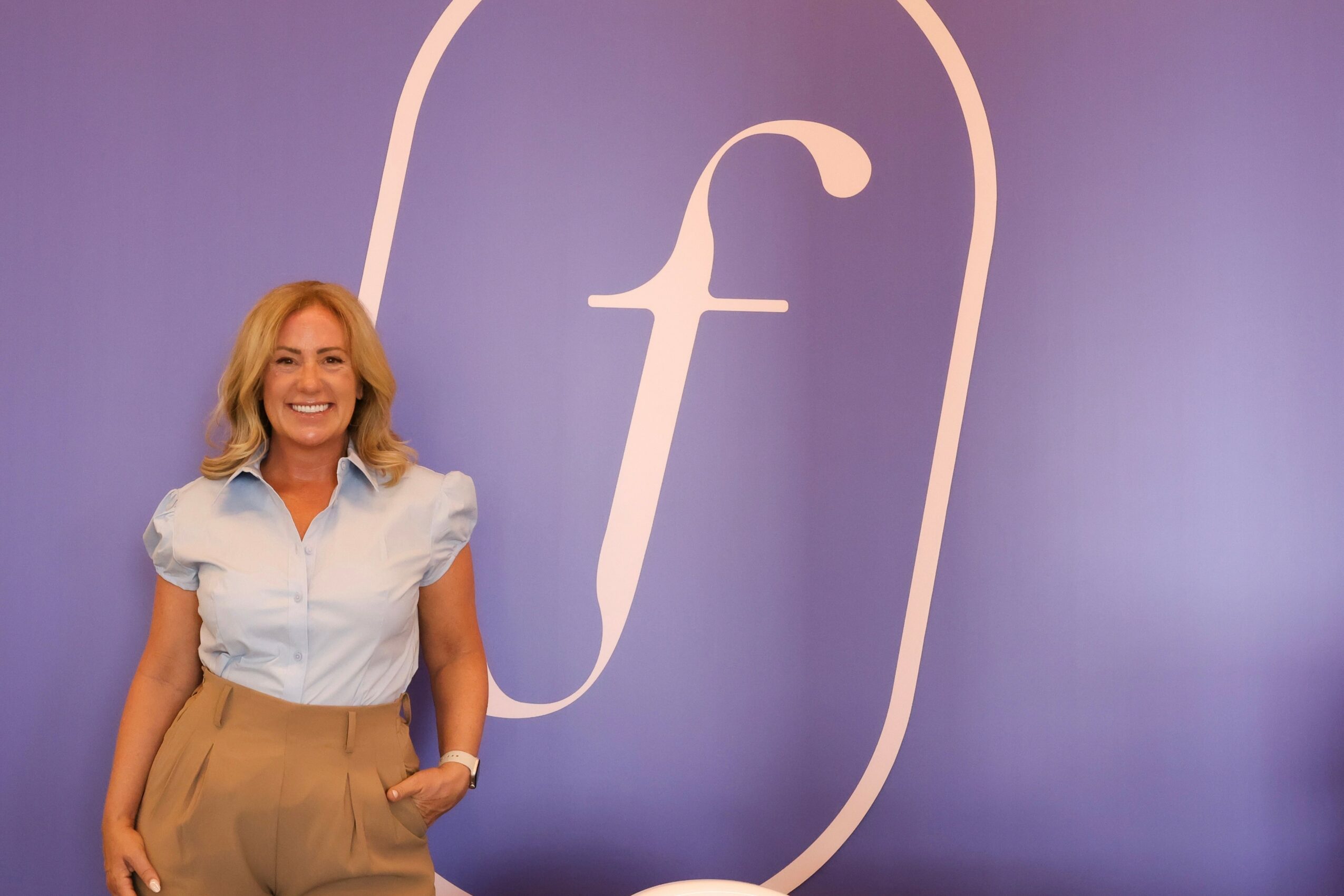 Woman with blonde hair smiling, standing in front of a purple background with a large white letter 'f' inside a rounded rectangle.