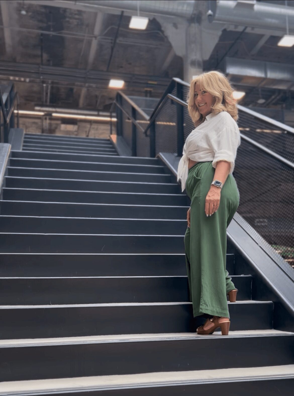 Woman with blonde hair in white blouse and green pants standing on stairs in industrial setting.