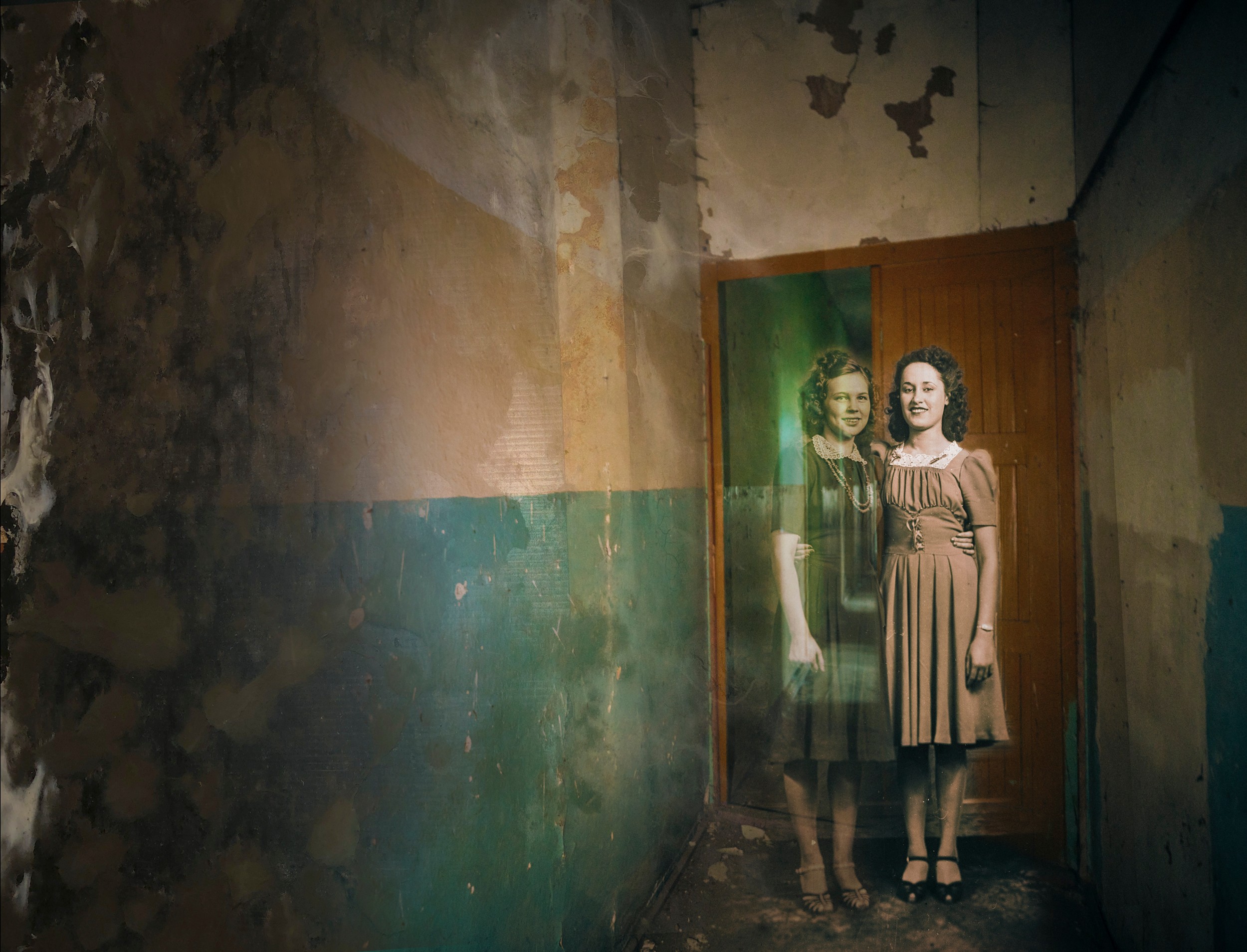 Two women stand in a doorway, with a mirror reflecting their images, in a dilapidated room with peeling walls.