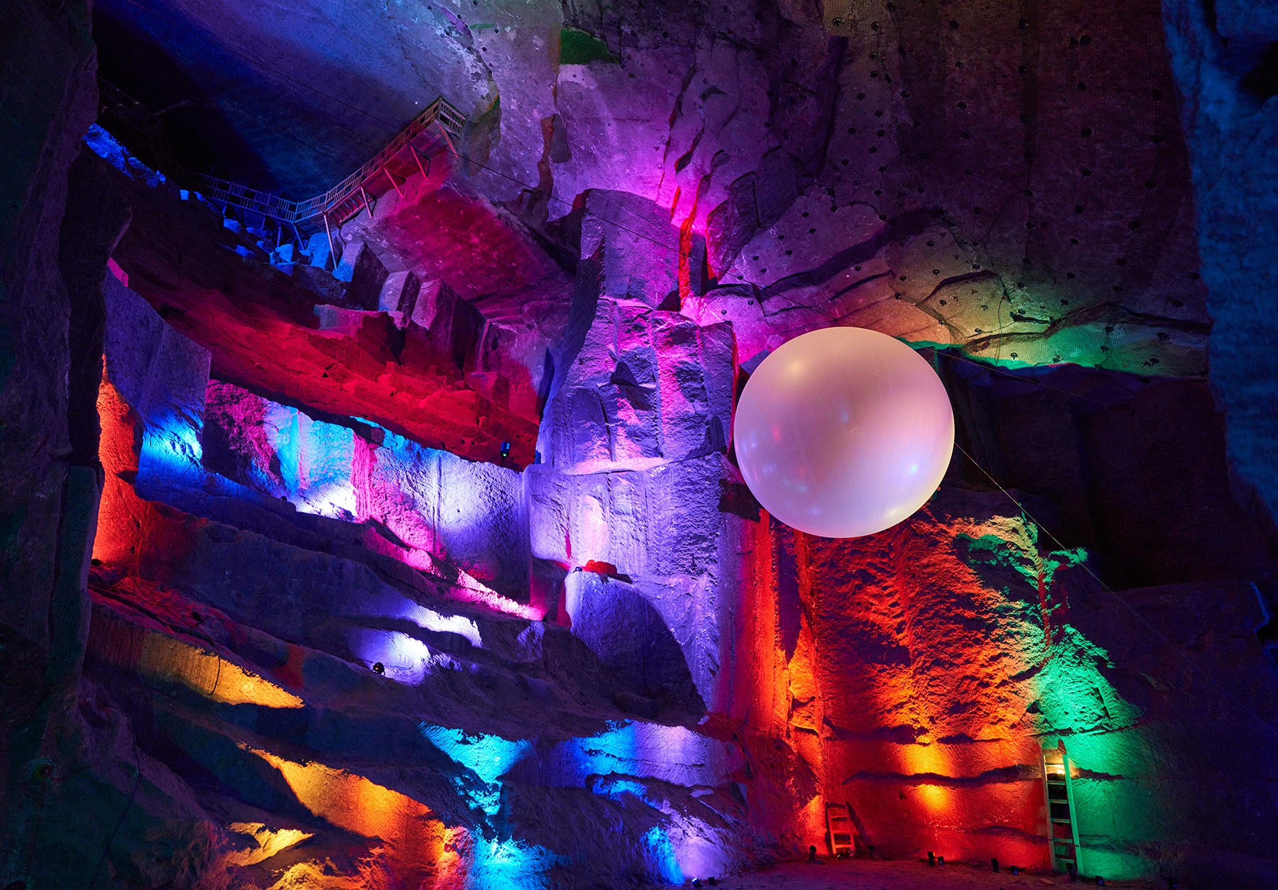 Colorful illuminated cave with a large spherical object hanging from the ceiling, vibrant rainbow lighting on rock formations.