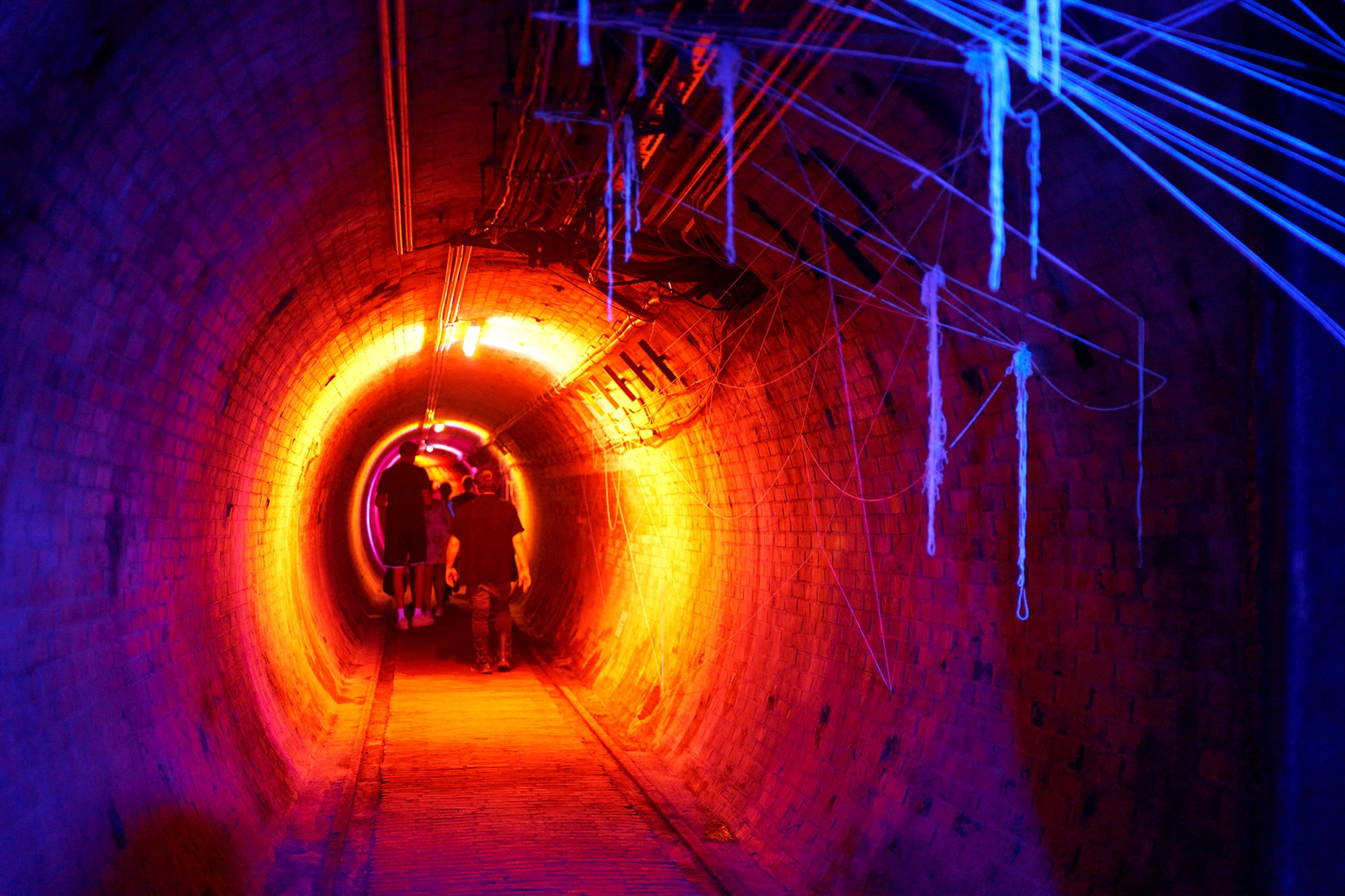 People walking through a tunnel illuminated with vibrant orange, blue, and purple lights, with glowing lines hanging from the ceiling.